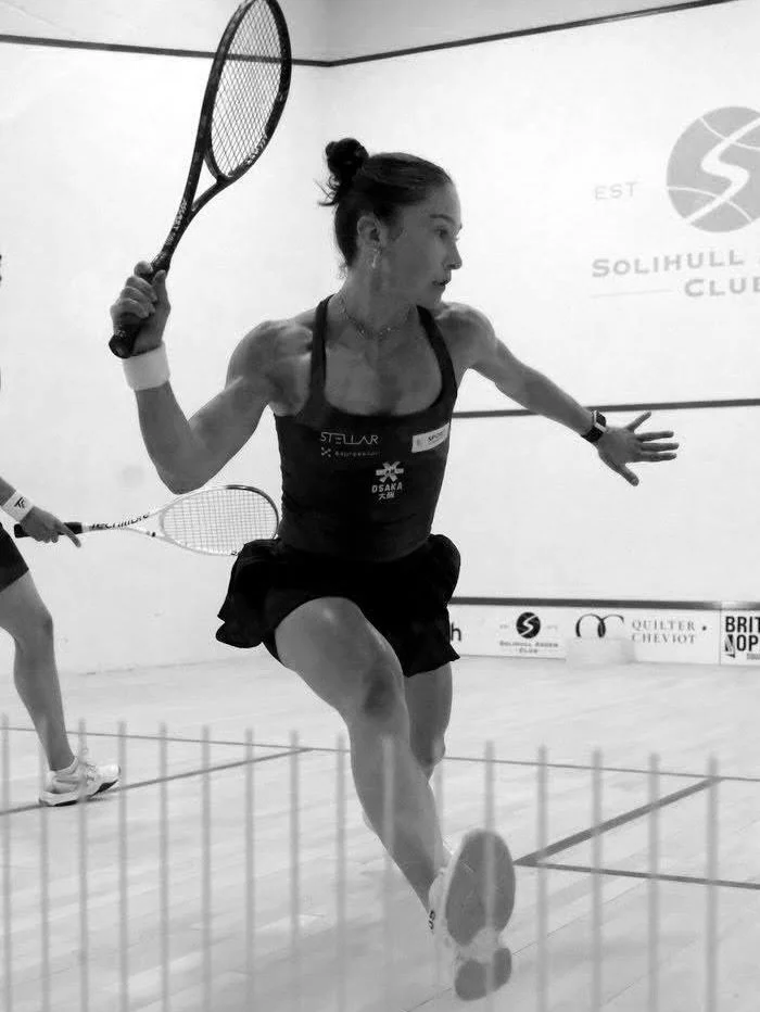 A woman playing squash on an indoor court, holding a racket with her right hand and extending her left arm, dressed in athletic wear.
