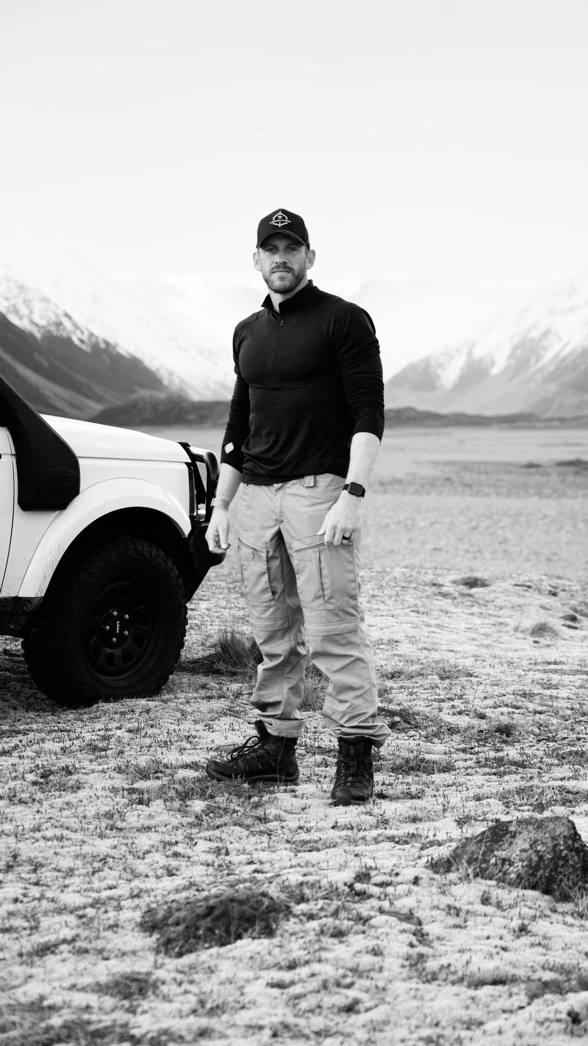 Man standing outdoors in a rugged landscape with mountains in the background, positioned next to a vehicle.