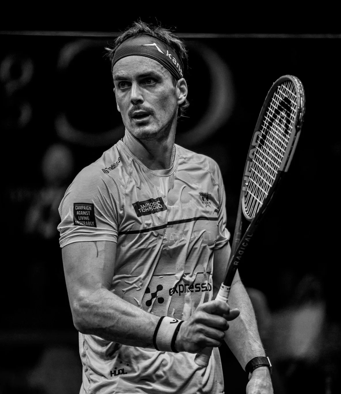 Paul Coll holding a squash racket during a match, wearing sportswear, a headband, and a wristband, captured in black and white.