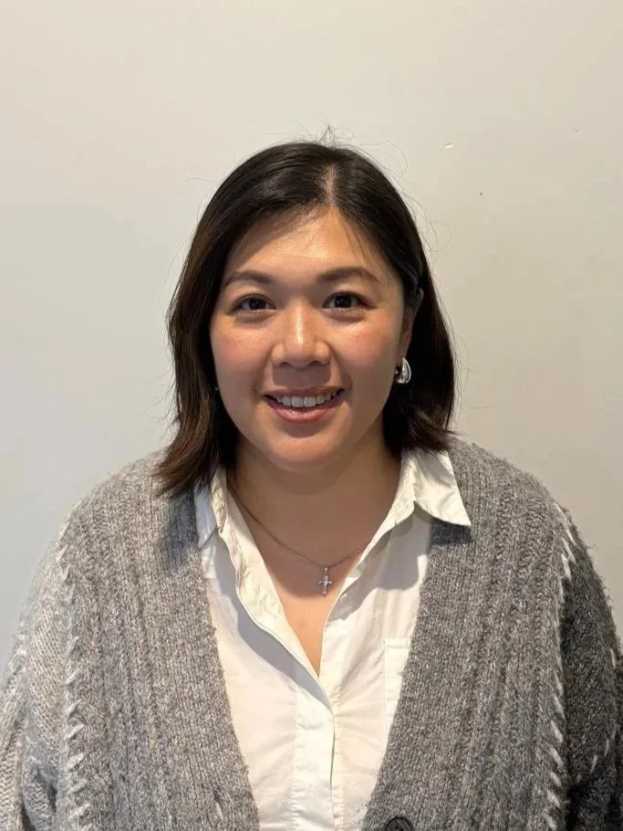 A smiling woman with shoulder-length dark hair, wearing a white shirt, a gray knitted cardigan, and a cross necklace, standing in front of a plain light-colored wall.