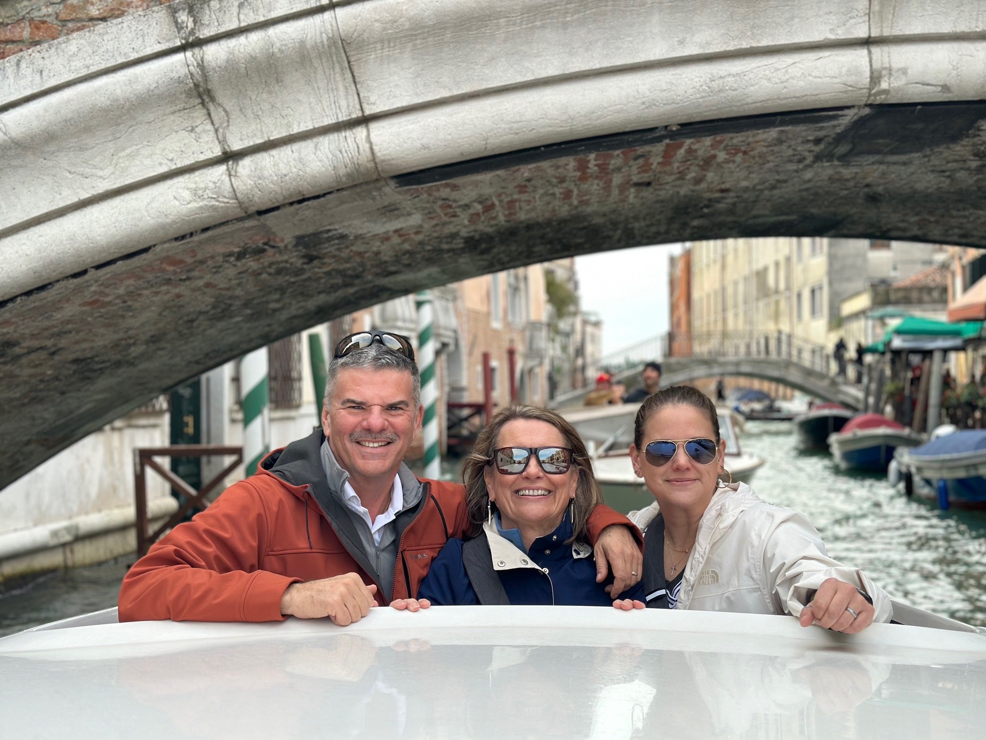 Four people smiling on a boat gondola ride in Venice, Italy, with historic buildings and a canal in the background.