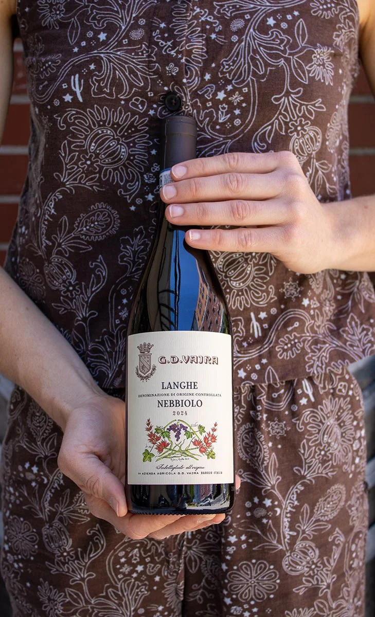 Person wearing a brown shirt with white floral and paisley patterns holding a bottle of Nebbiolo wine from 2024.