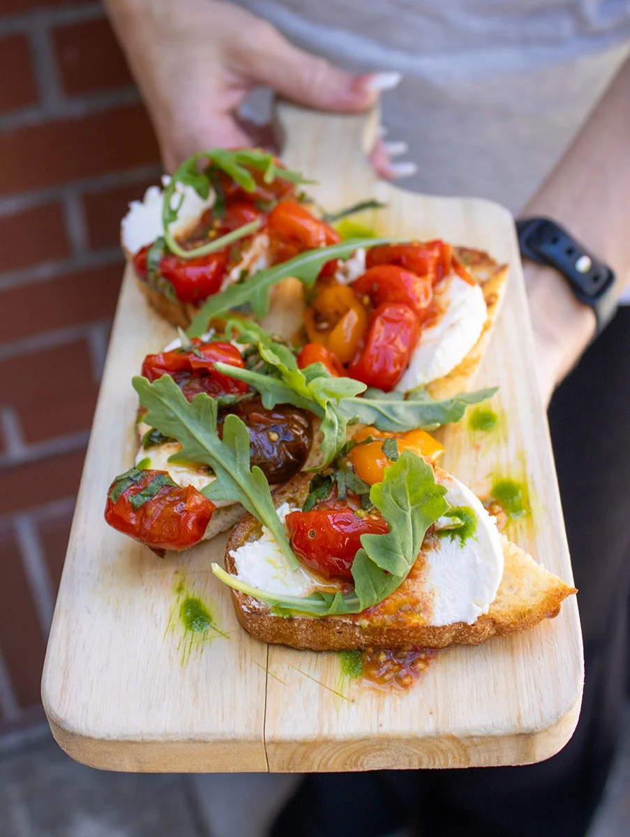Person holding a wooden serving board with three slices of bruschetta topped with cherry tomatoes, arugula, fresh mozzarella, and drizzled with green pesto oil.