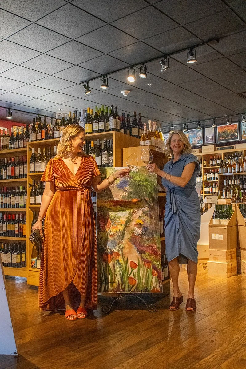Two women standing in a wine shop, holding wine glasses, standing beside a colorful painting of a garden scene with flowers and greenery.