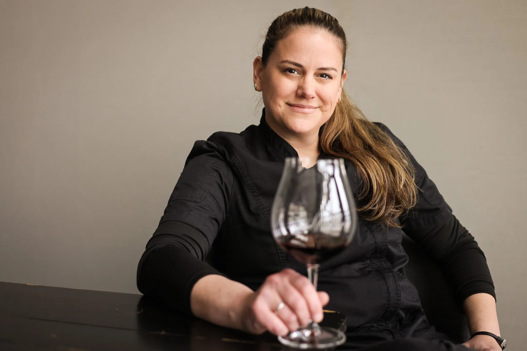 A woman with long, wavy hair sitting at a table with a glass of red wine in front of her, smiling at the camera, against a beige wall.