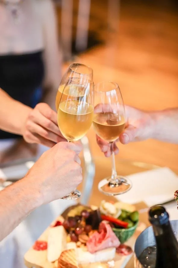 Three people clinking glasses of white wine during a celebration or gathering, with a cheese and meat platter on the table.