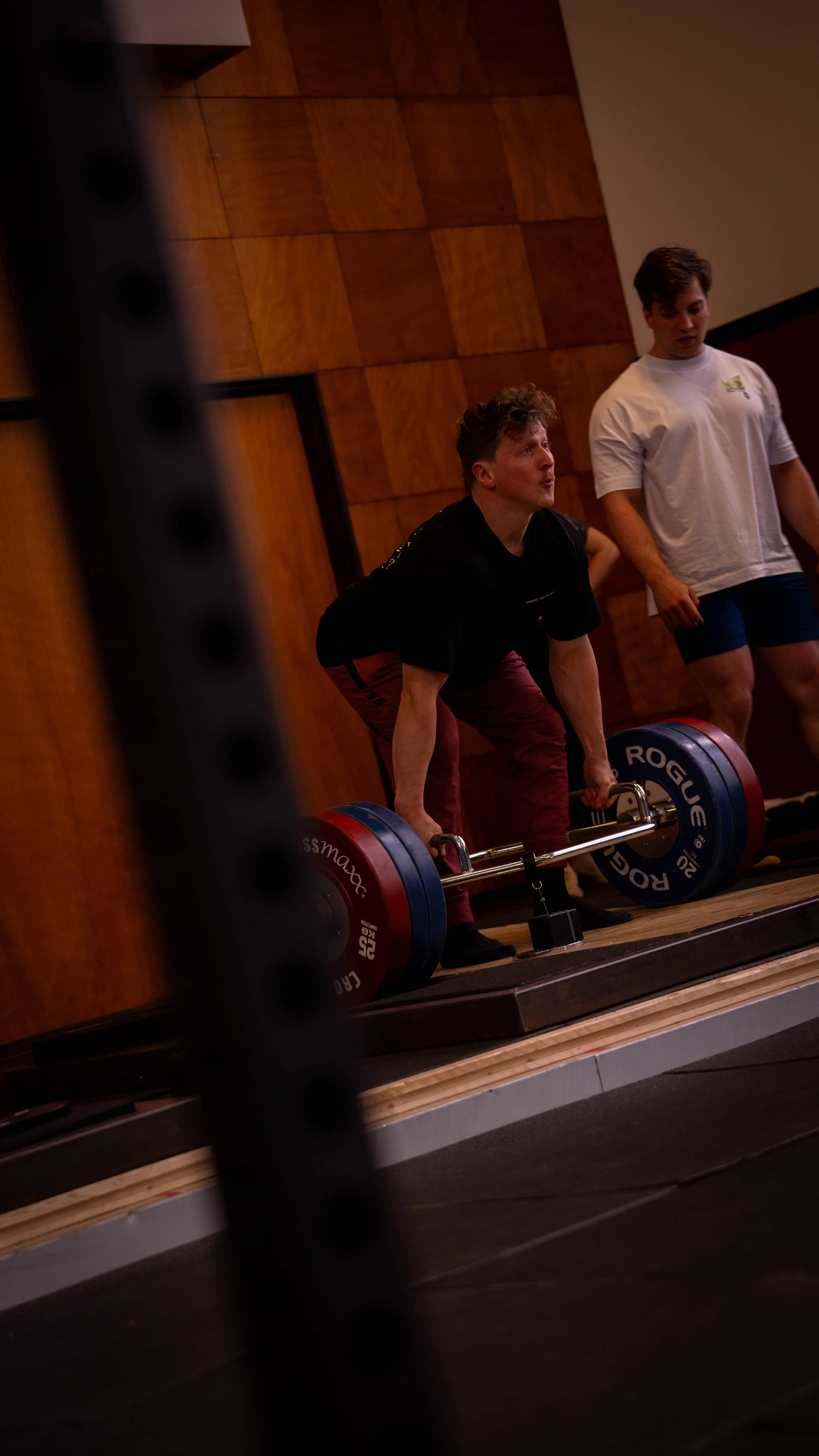 A person performing a deadlift exercise with a loaded barbell, wearing a black shirt and maroon pants, in a gym with two onlookers nearby.