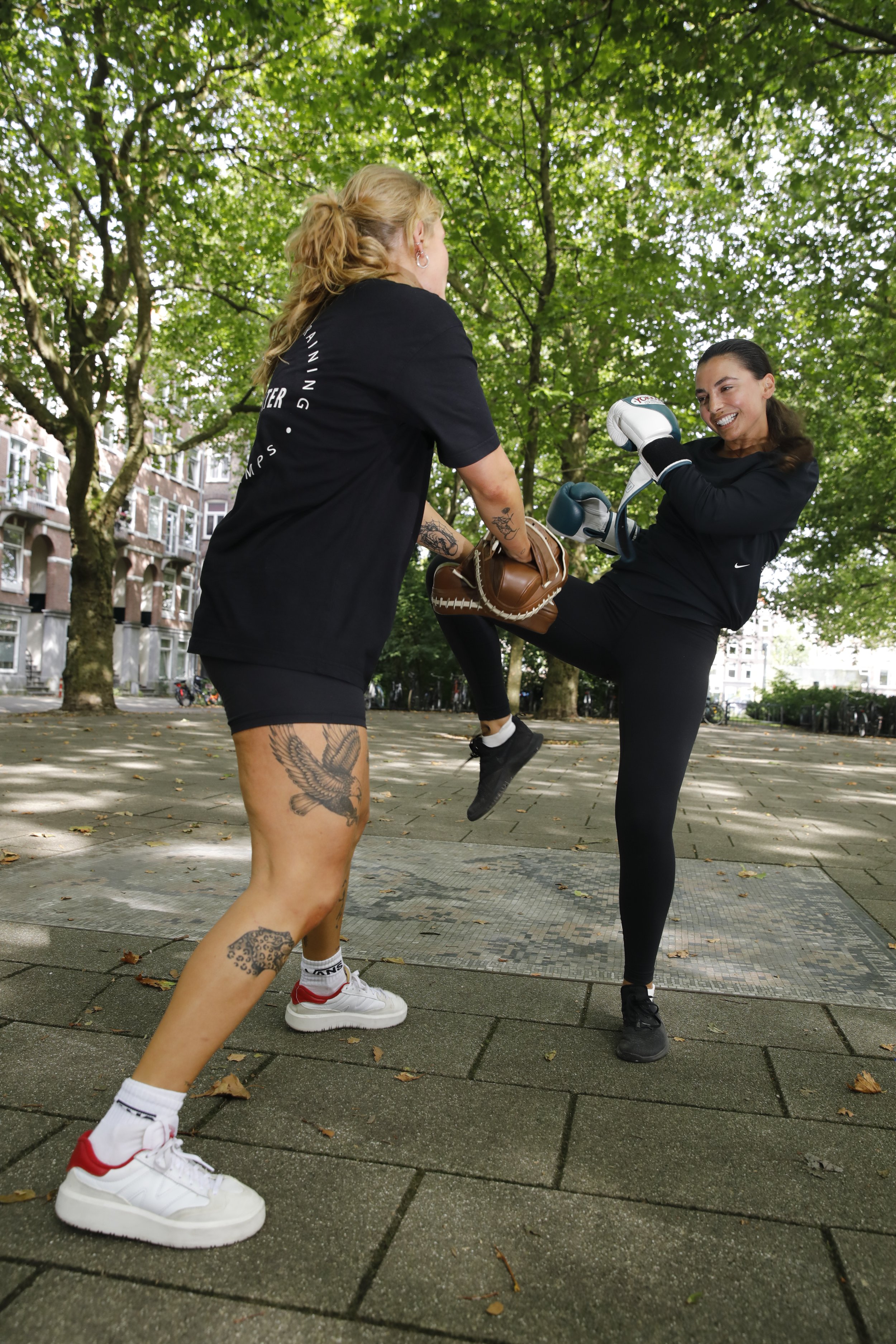 Two women practicing martial arts outdoors in a park, one in a black shirt and shorts with tattoos, the other in a black athletic outfit with boxing gloves, during daytime.