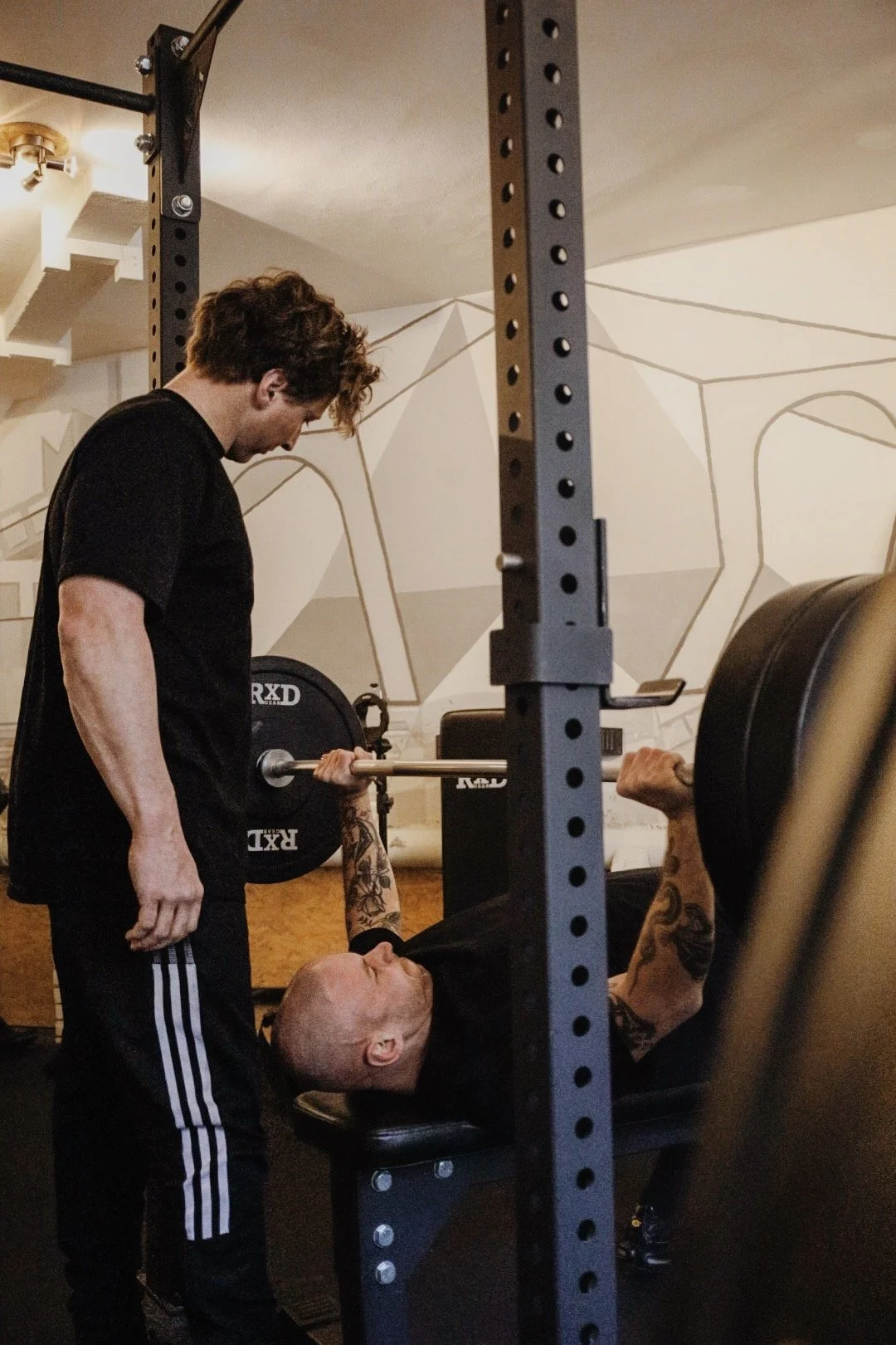 A man spotter assists another man during a bench press exercise at the gym, with barbell and weights.