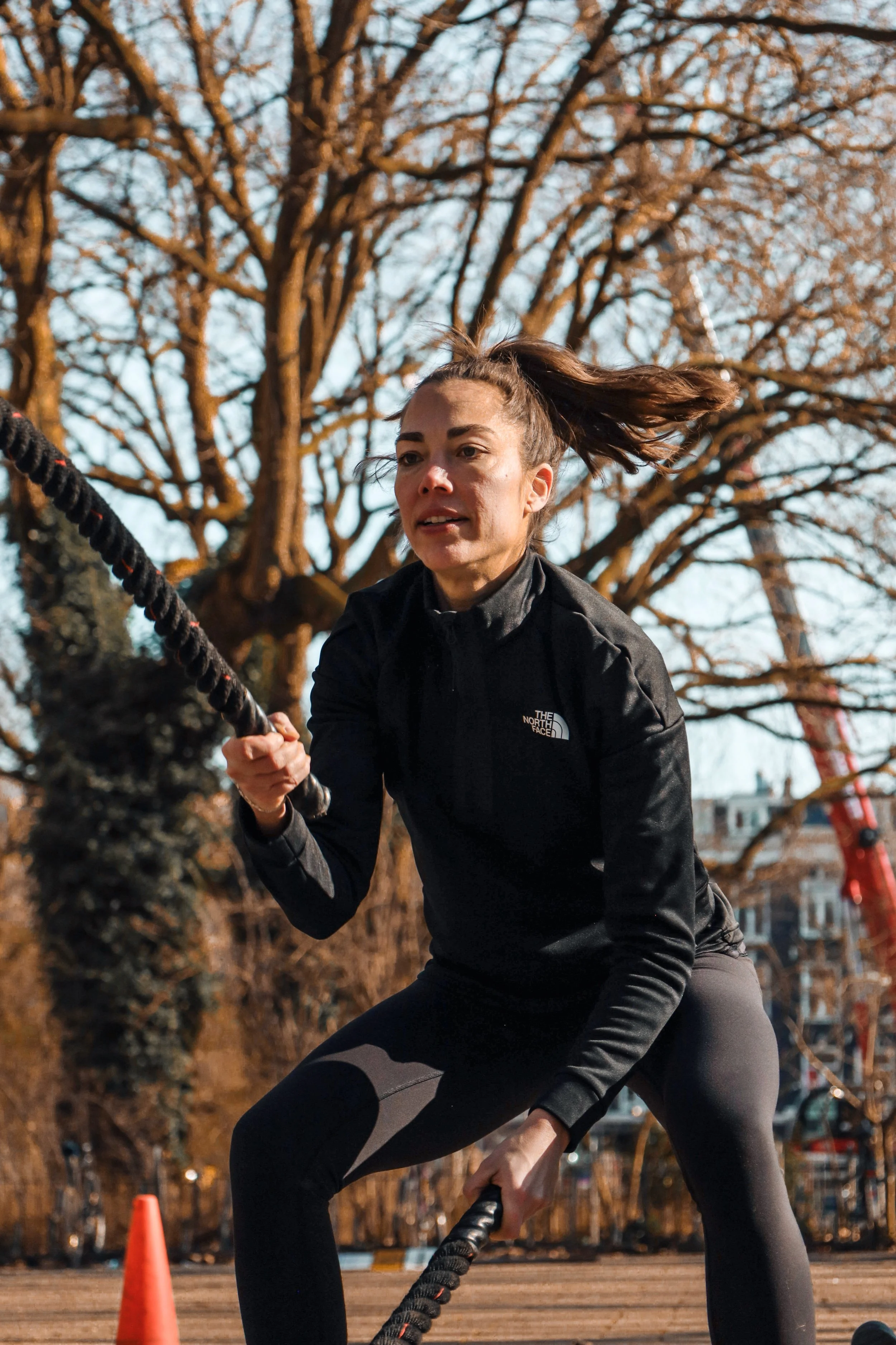 A woman in black athletic clothing, with her hair tied back, is pulling a rope outdoors during daytime. She appears to be in an intense workout or competition, with a large tree and a cityscape in the background.