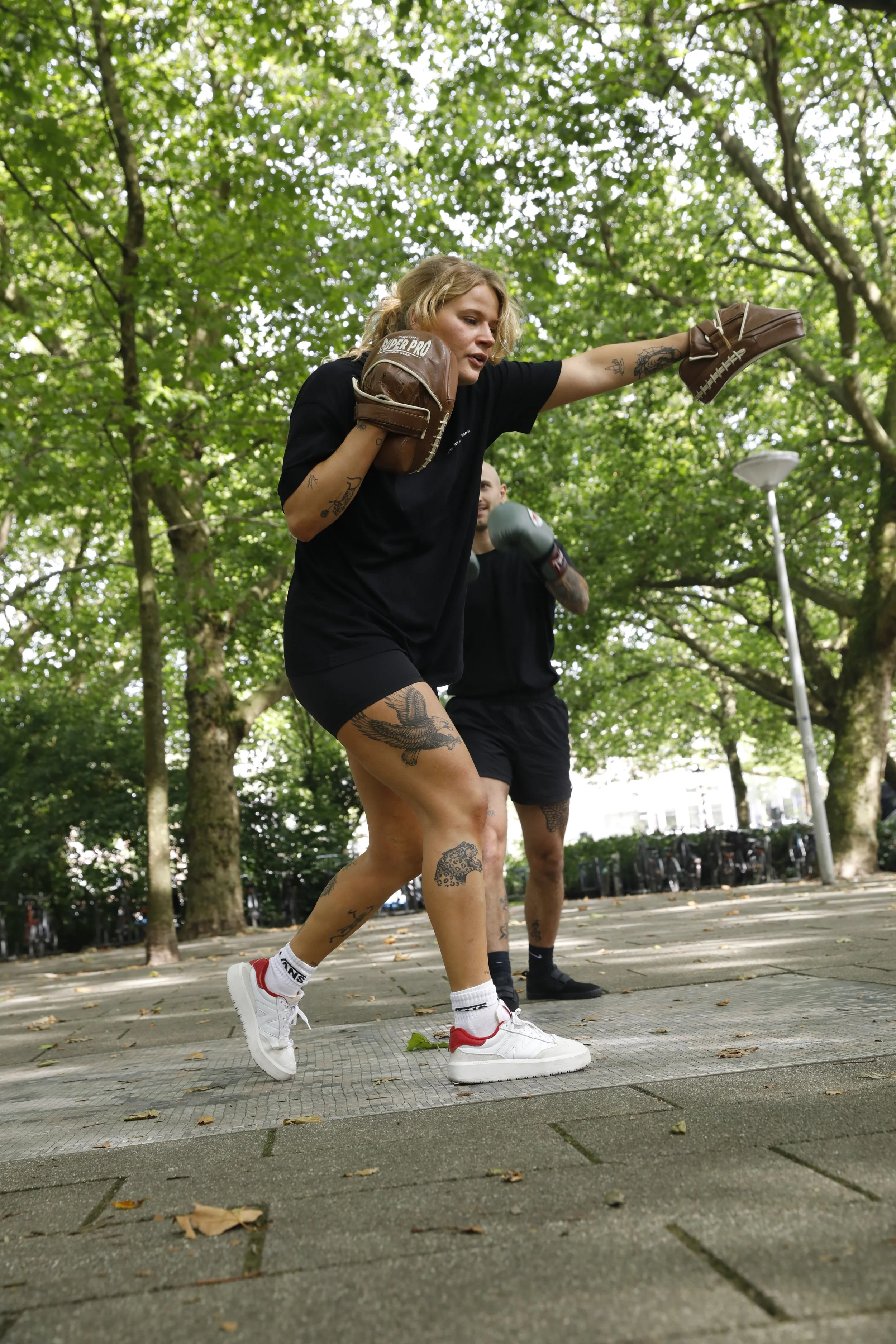 Two women practicing boxing outdoors in a park with green trees, one woman in front throwing a punch while the other is behind her with boxing gloves, both dressed in black athletic wear and white sneakers.
