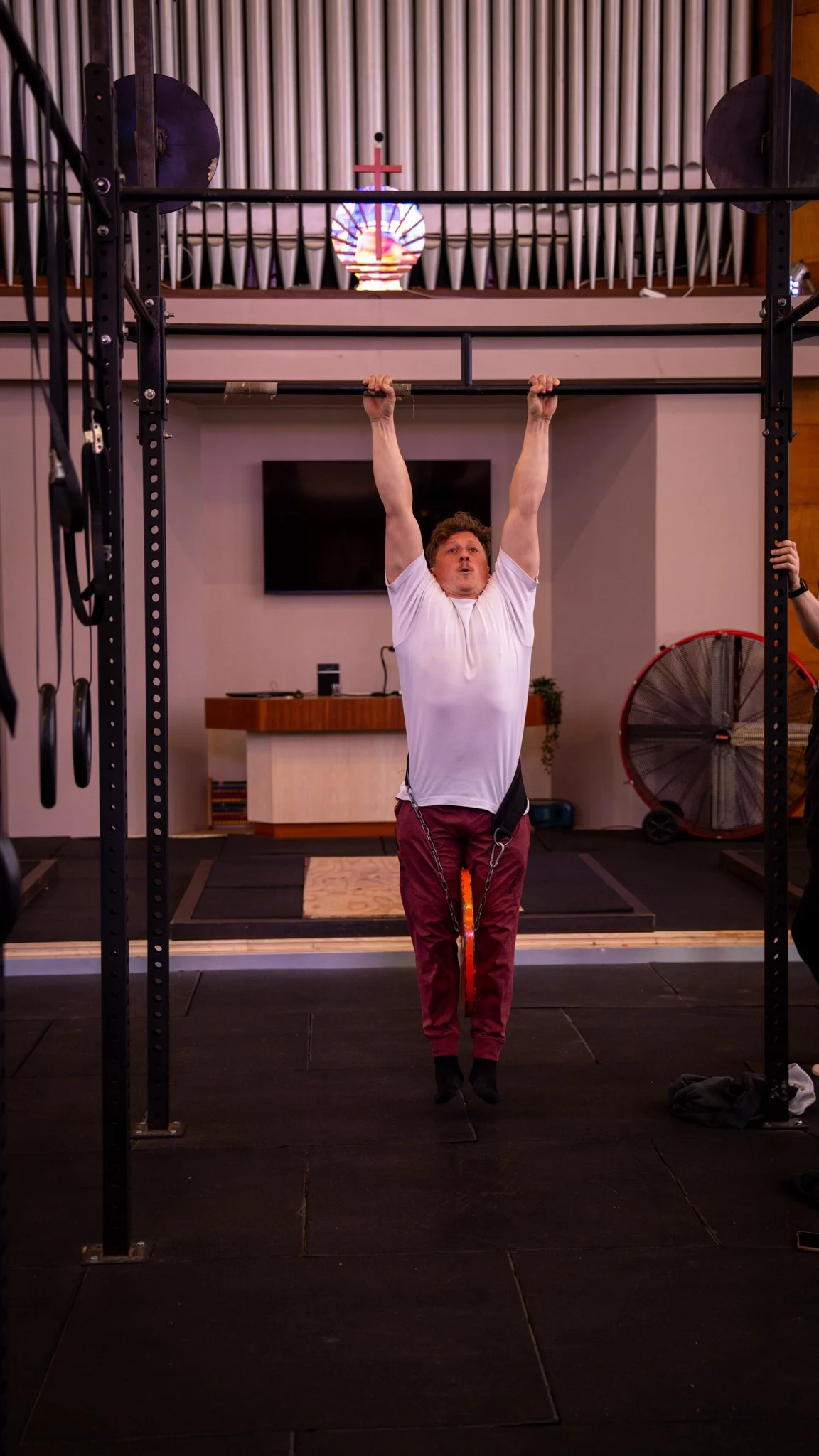 A man in a white shirt and maroon pants lifting a barbell overhead in a gym. The gym has black flooring, a TV on the wall, and exercise equipment visible.