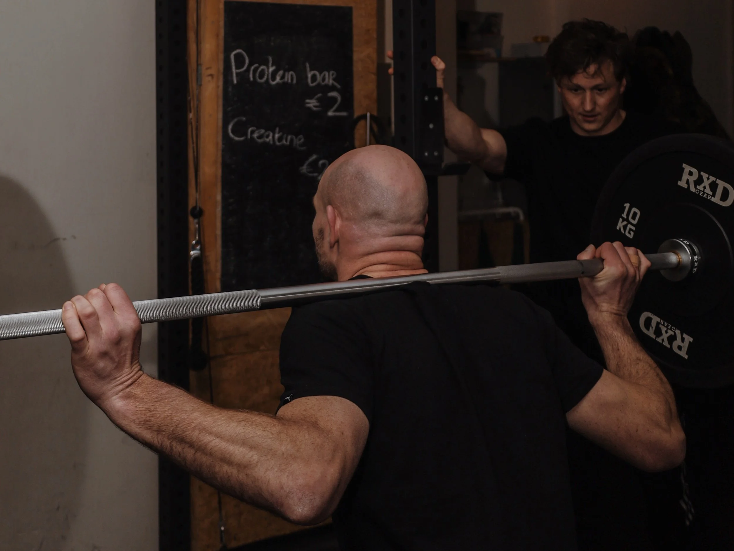 A man lifting a barbell with weights in a gym, with a trainer standing behind him.