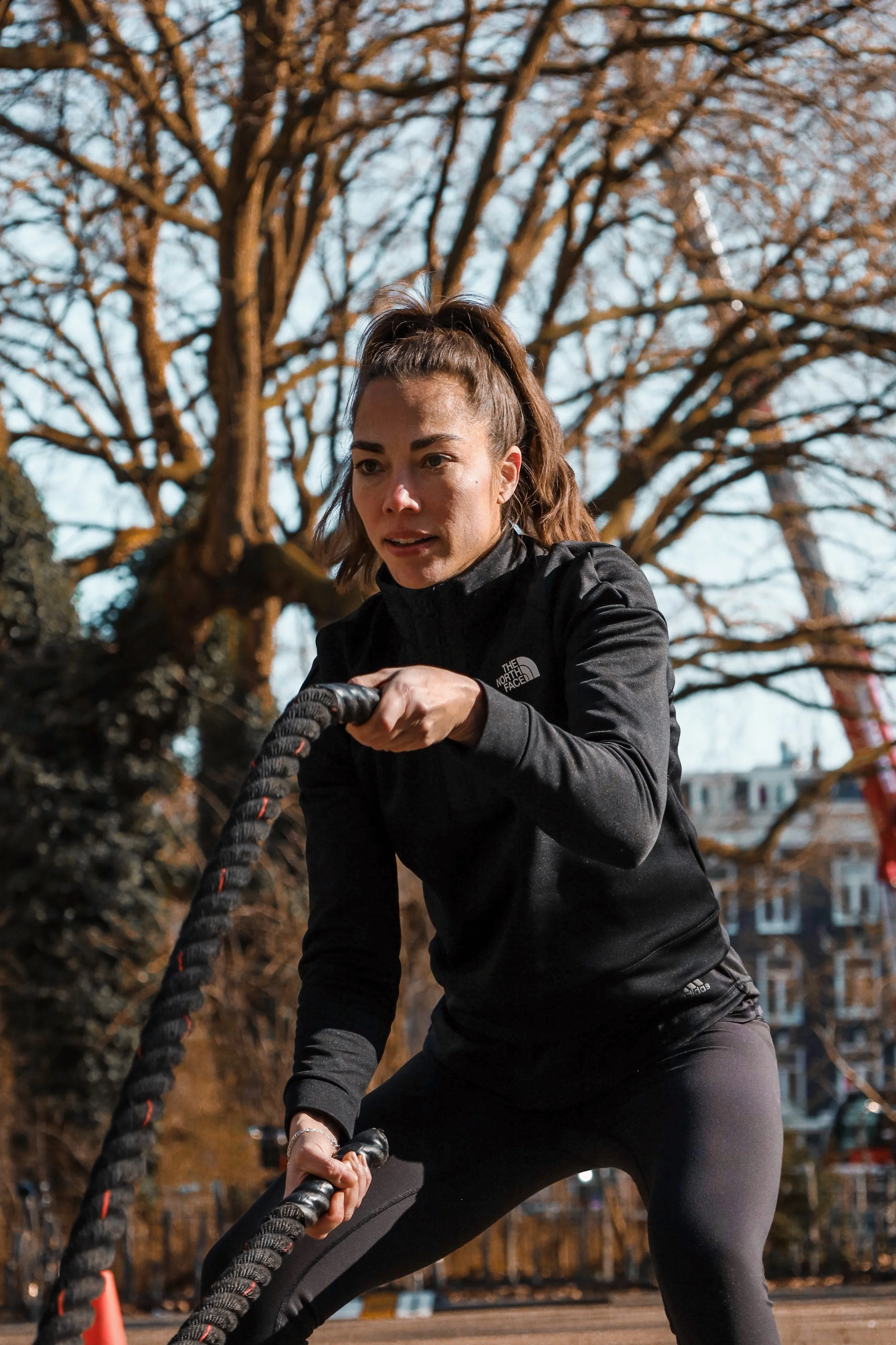 Woman in black sportswear pulling battle ropes outdoors with trees and buildings in the background.
