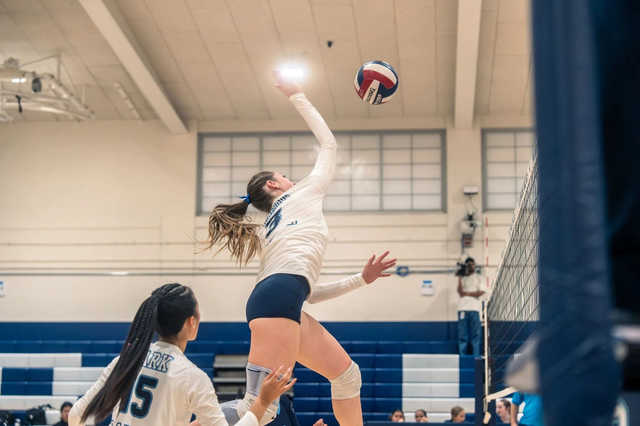 A female volleyball player jumps near the net to hit the ball during a match, with her teammates watching from below in a gymnasium.