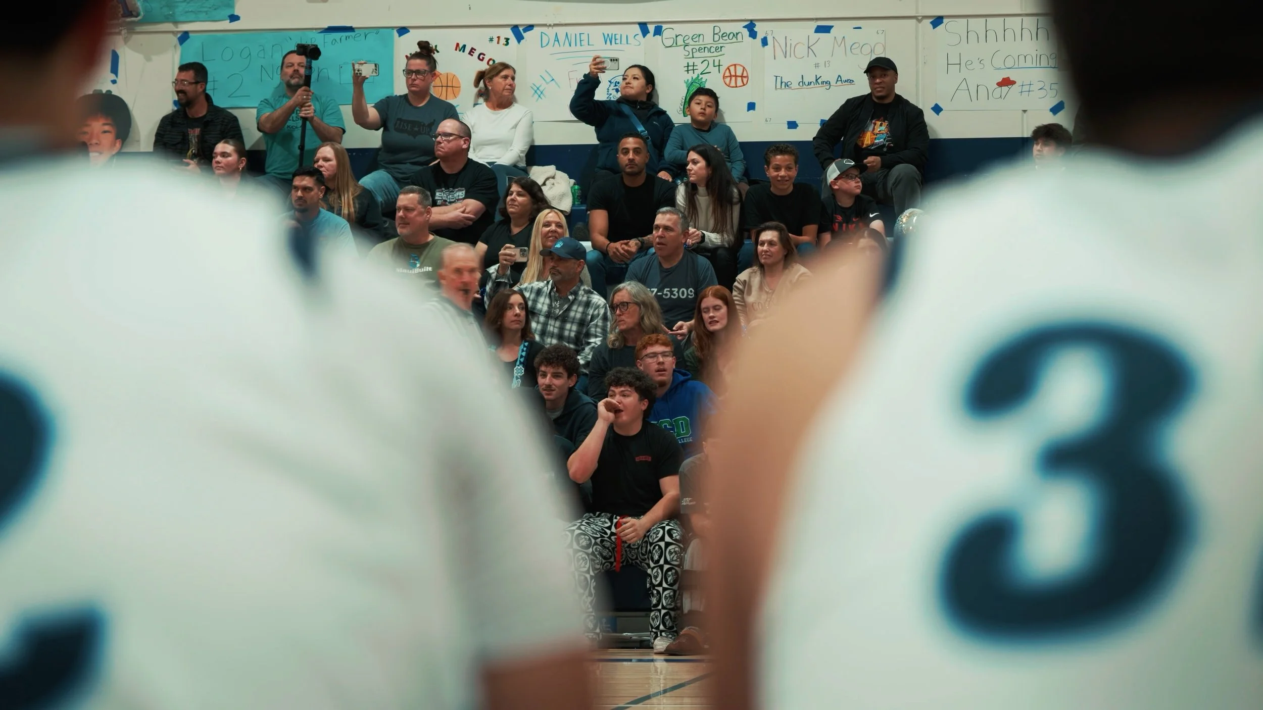 View of a crowded gymnasium with spectators seated in bleachers, some holding phones, and signs with names and numbers on the wall behind them, seen through large, out-of-focus volleyballs.