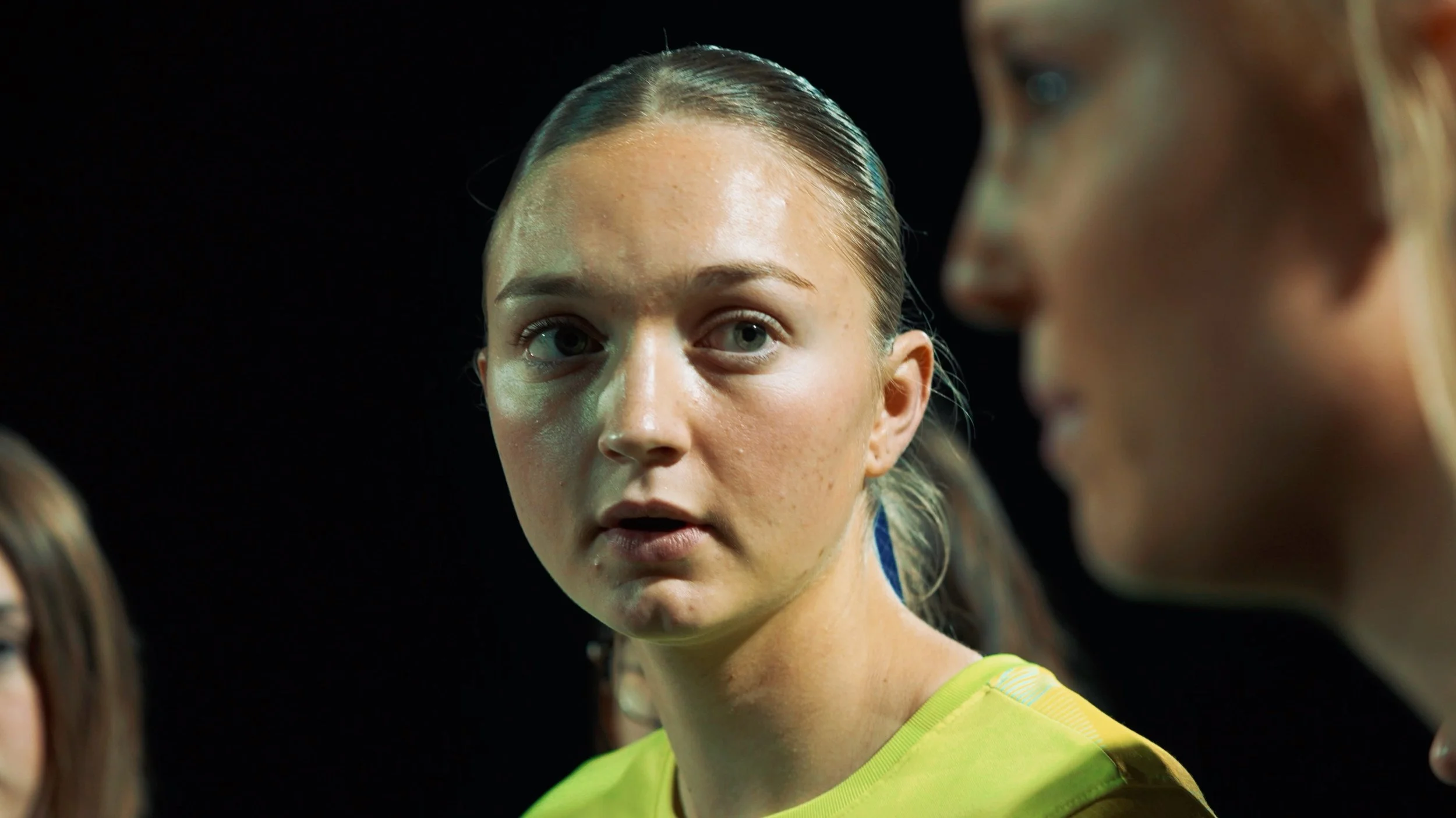 Close-up of a young woman with blonde hair slicked back, wearing a yellow sports jersey, looking attentively at someone outside the frame.