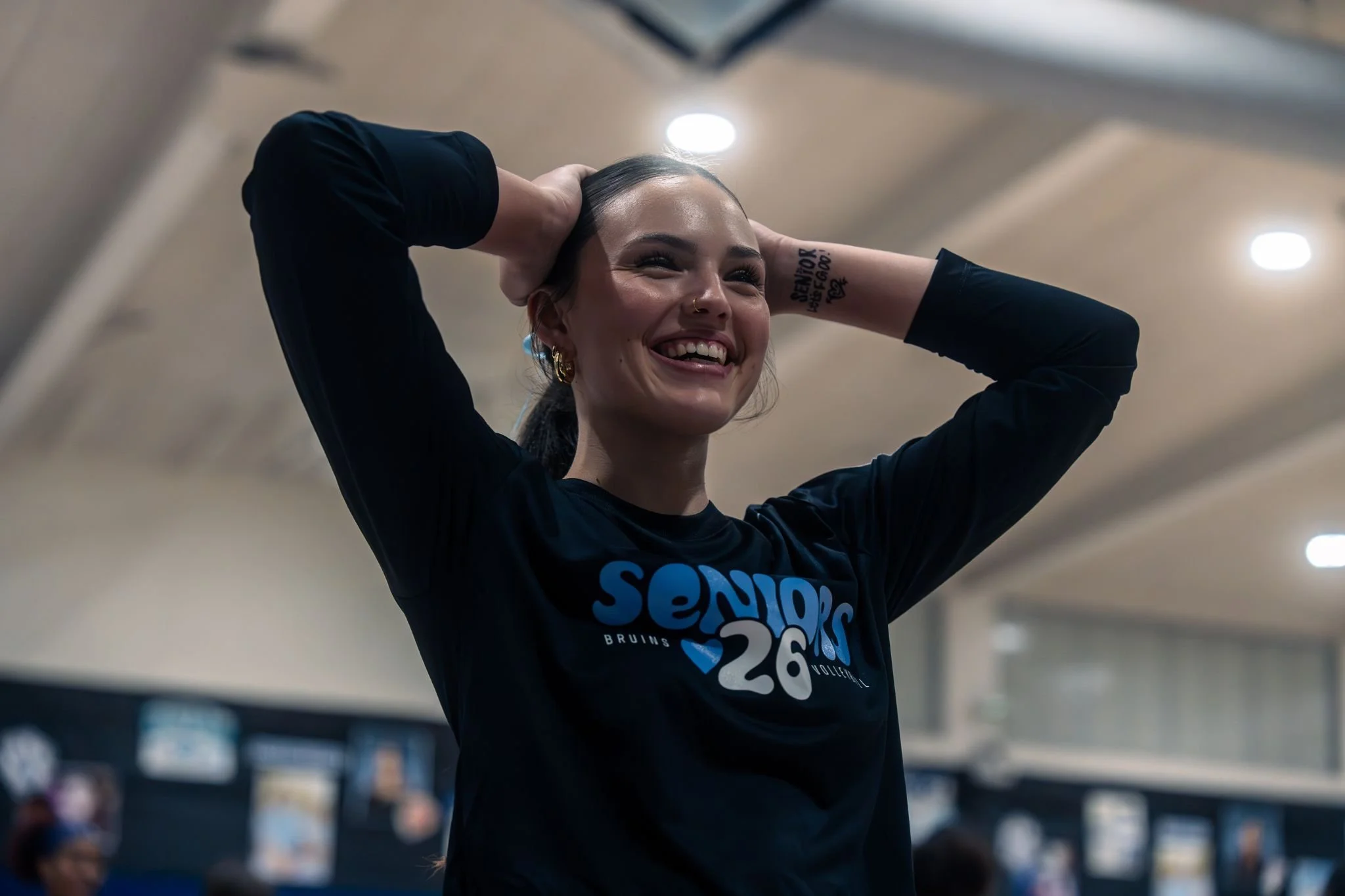 A smiling woman with dark hair pulled back, wearing a black long-sleeve shirt with blue and white text, holding her hands behind her head in a public indoor setting with bright ceiling lights.