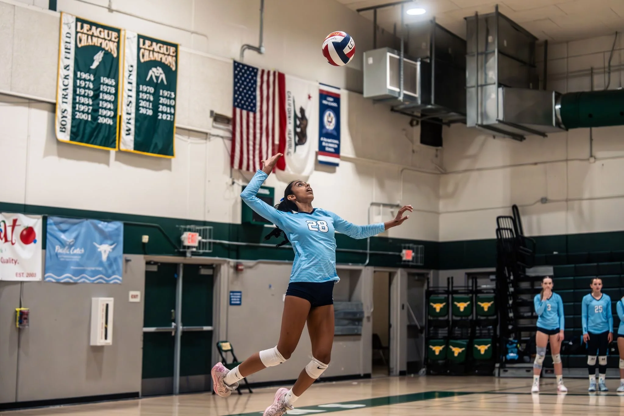 A volleyball player wearing a light blue jersey with the number 28 jumps to hit the ball during an indoor match. Other players and banners are visible in the background.