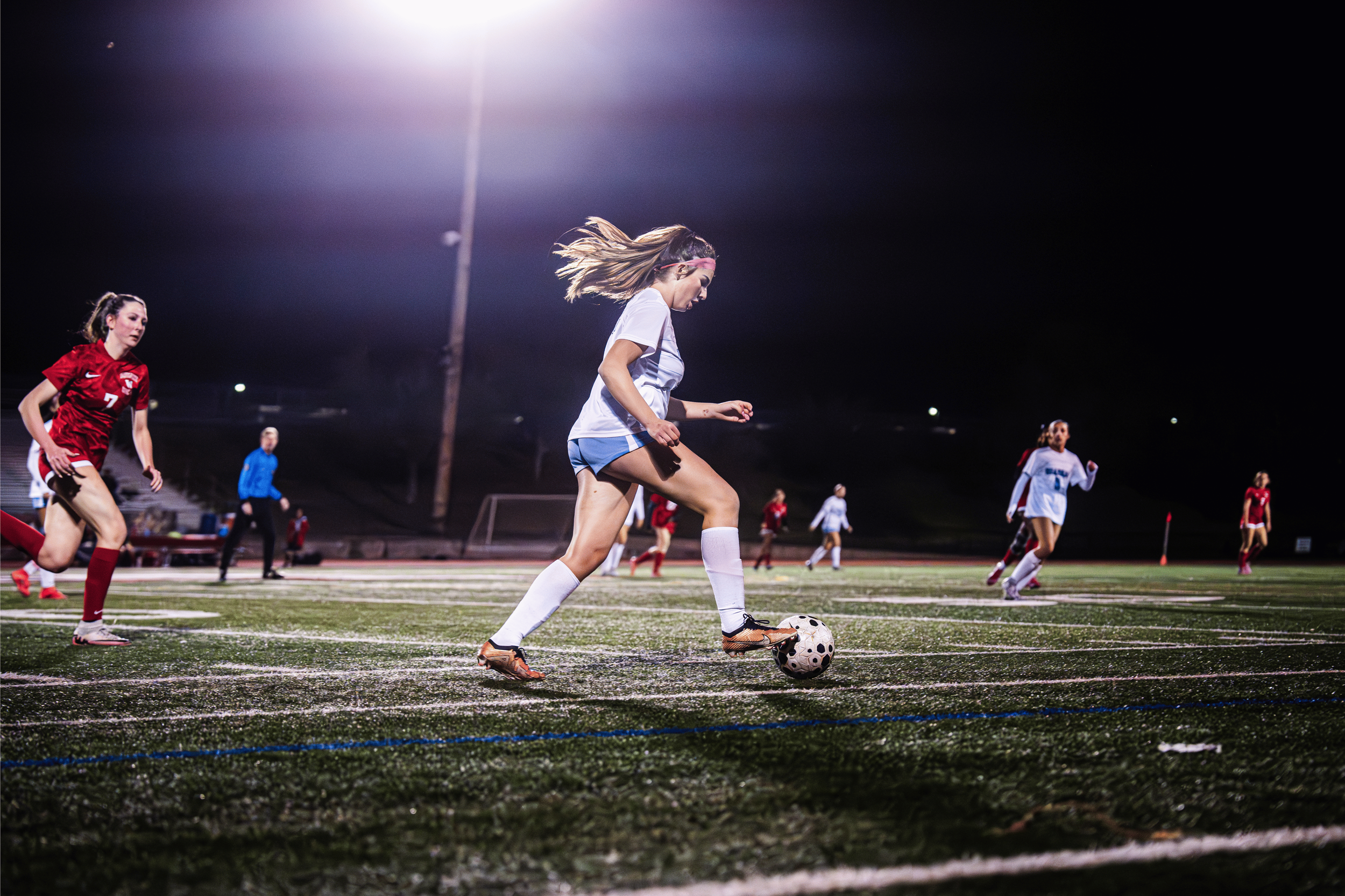 A young girl in a white sports uniform with blue shorts and a pink headband playing soccer on a field under bright stadium lights at night, with other players and a referee in the background.