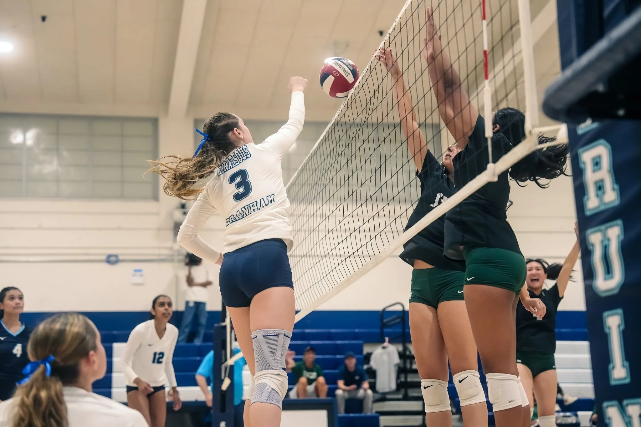 Volleyball players in a gym, one in white with the number 3, blocking a ball at the net against opponents in black and green uniforms.