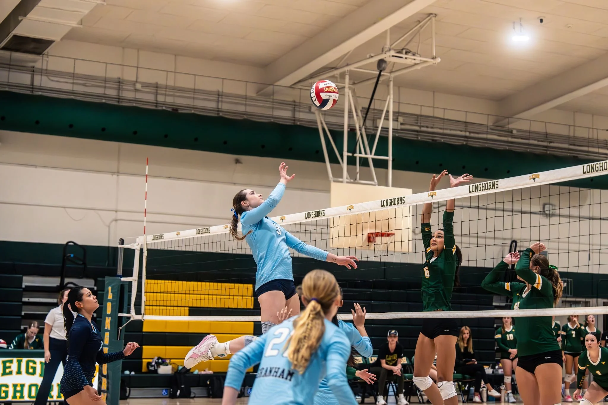 A volleyball player in a light blue uniform jumps to hit the ball over the net as opponents in green uniforms attempt to block. The scene takes place indoors with spectators watching.