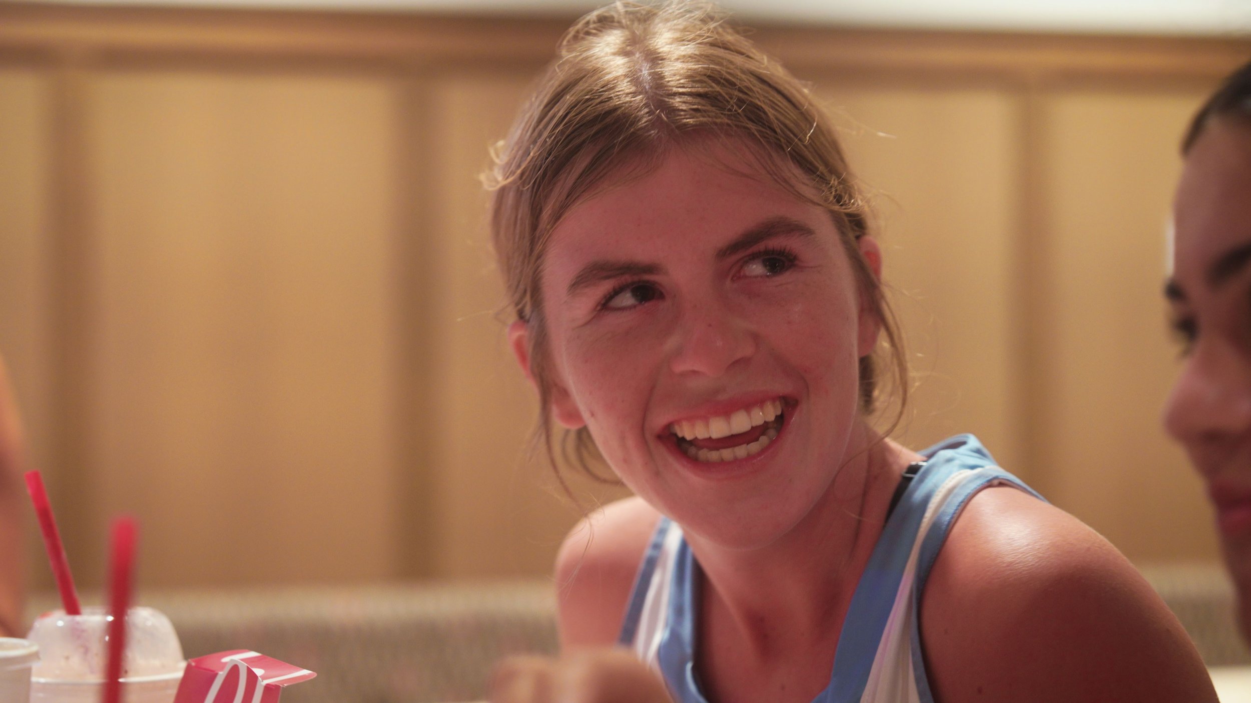 A young woman with blonde hair smiling and looking to her right, wearing a sports tank top, in a setting with a warm wooden background.