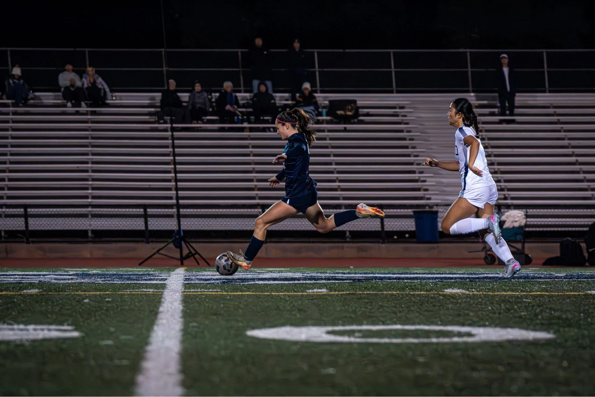 Two female soccer players in dark and white uniforms running on a field at night, with spectators sitting on bleachers in the background.
