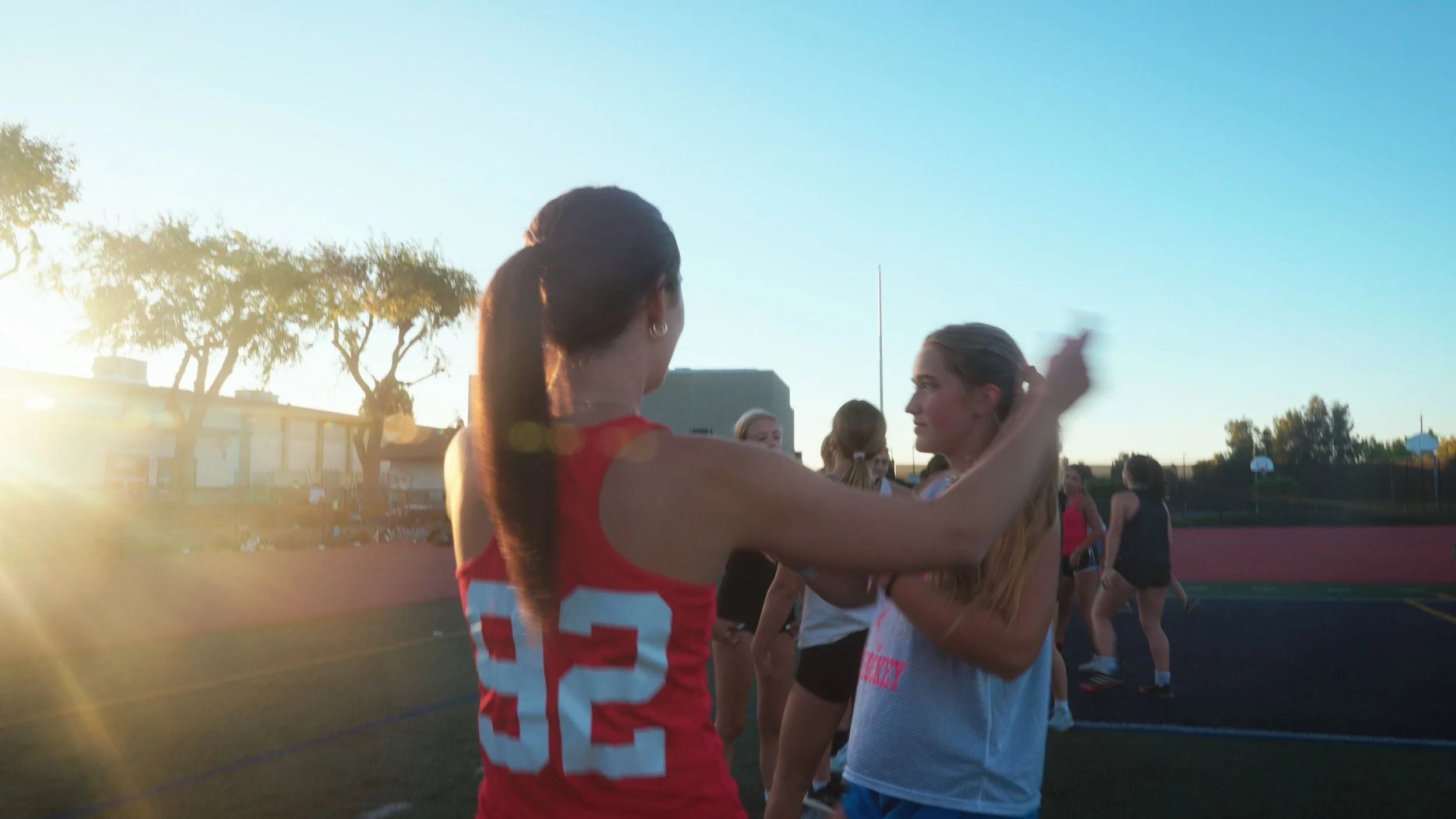 A group of young women, some in sports uniforms, interacting and smiling on a track field during sunset.