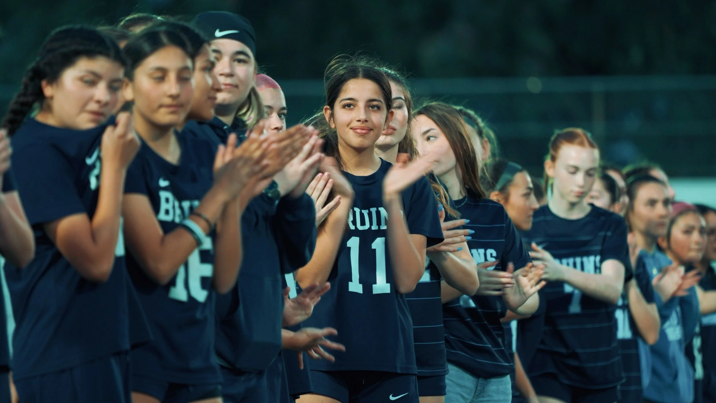 A group of young female athletes in matching dark blue sports jerseys, standing in a line, some clapping, on a sports field during evening.