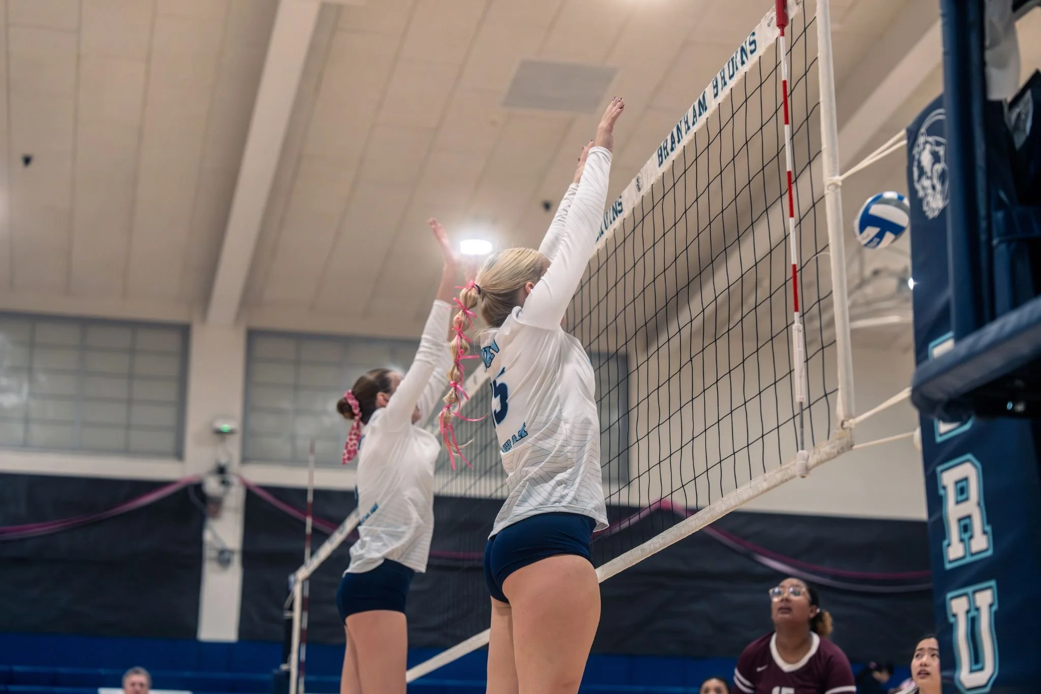 Women playing volleyball at indoor court, jumping near net, wearing white uniforms.