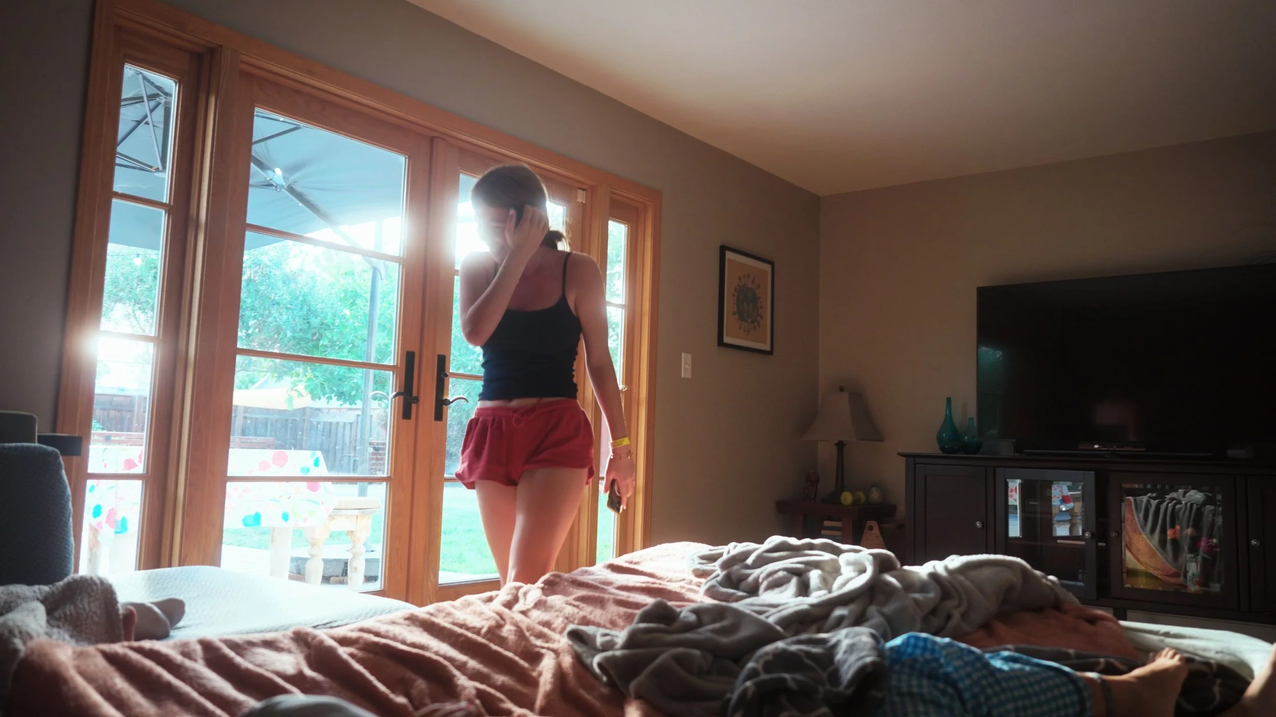 A woman in a black tank top and red shorts talking on her cell phone inside a living room, standing near sliding glass doors that lead to a backyard with an umbrella and trees.