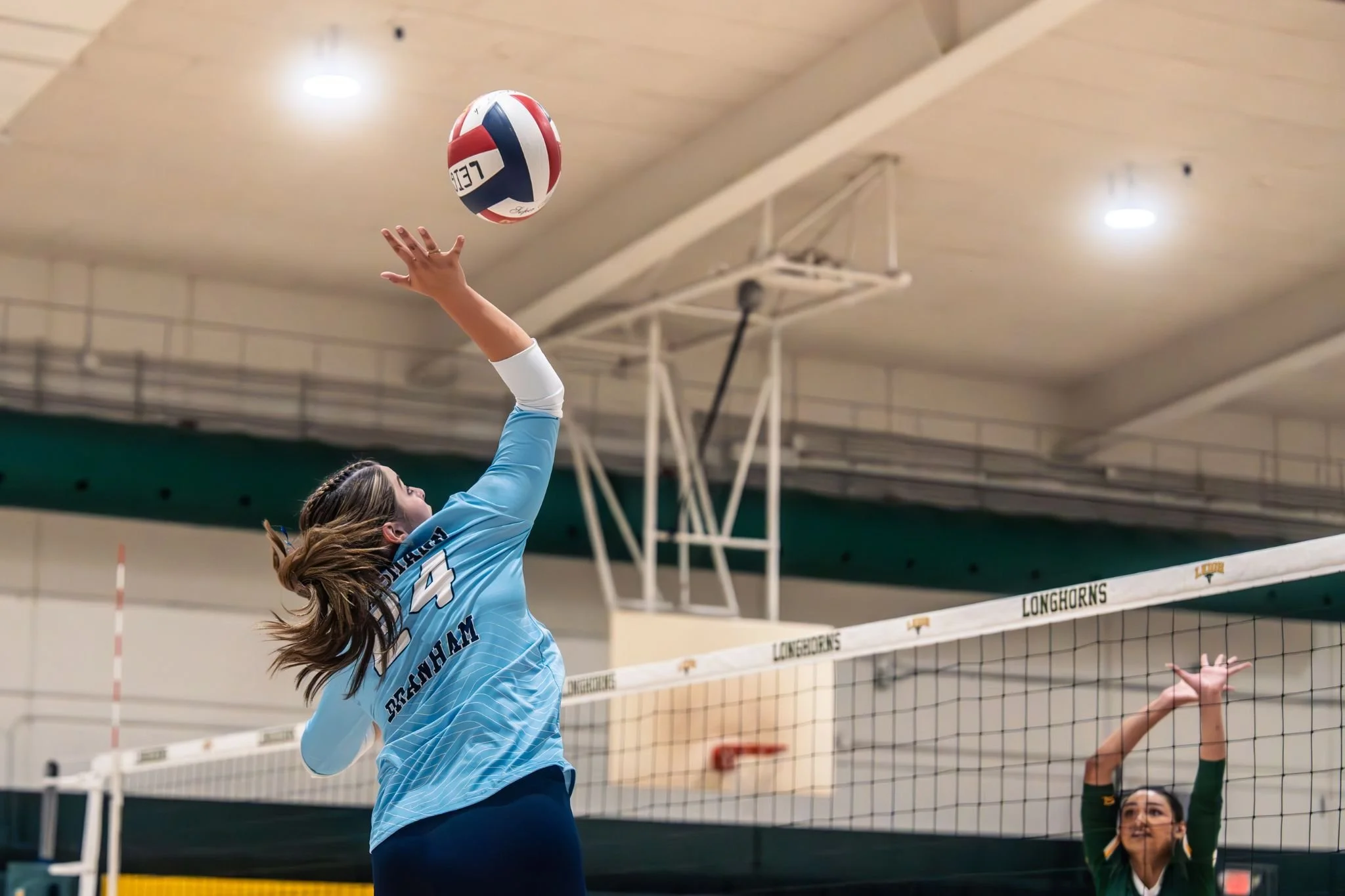 A female volleyball player in a light blue jersey jumps to hit the volleyball over the net during a game in an indoor gym.