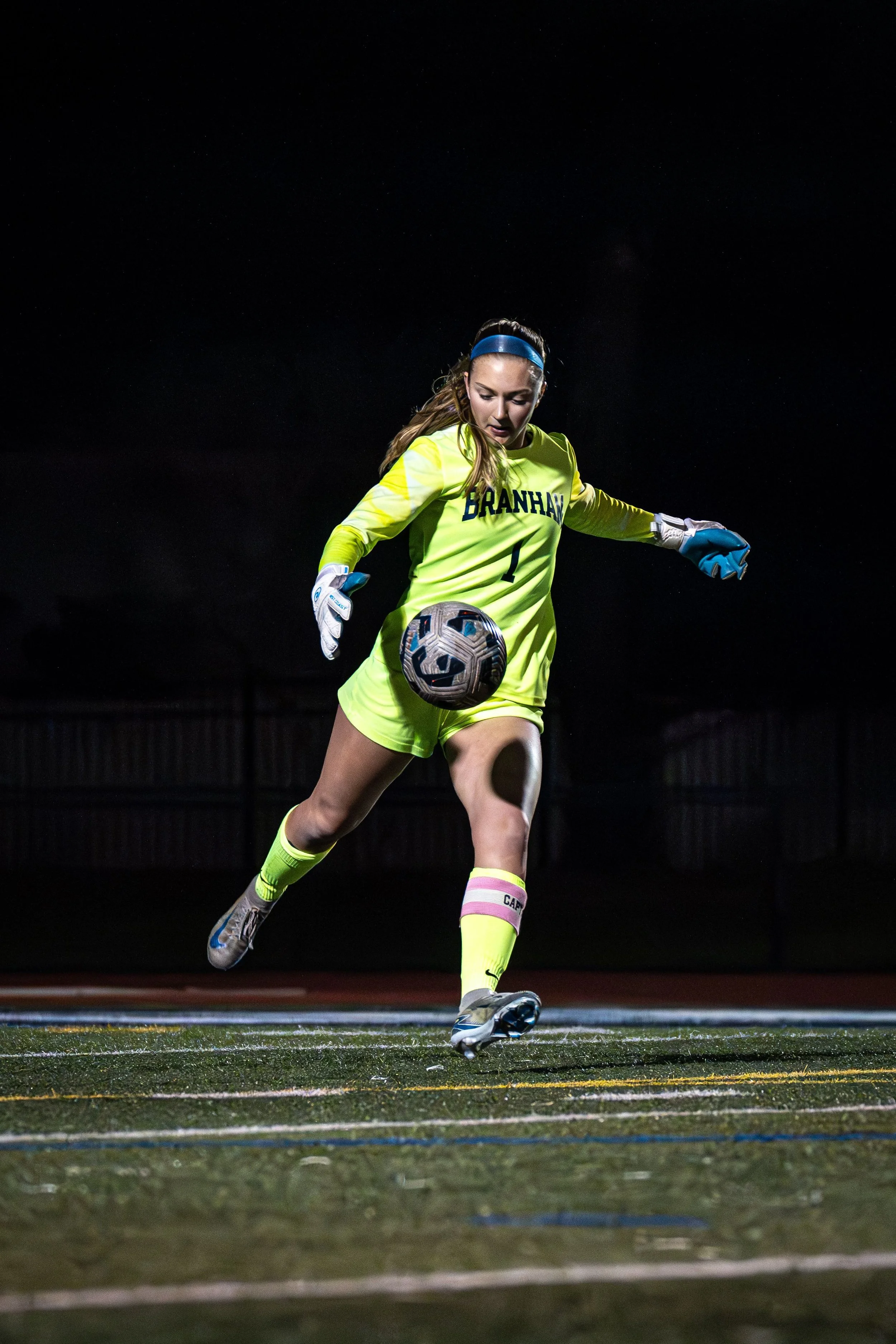 A female soccer goalkeeper in neon yellow uniform with the word 'BRANHAM' on her jersey, wearing a blue headband, positioning herself to catch a soccer ball on a night game.