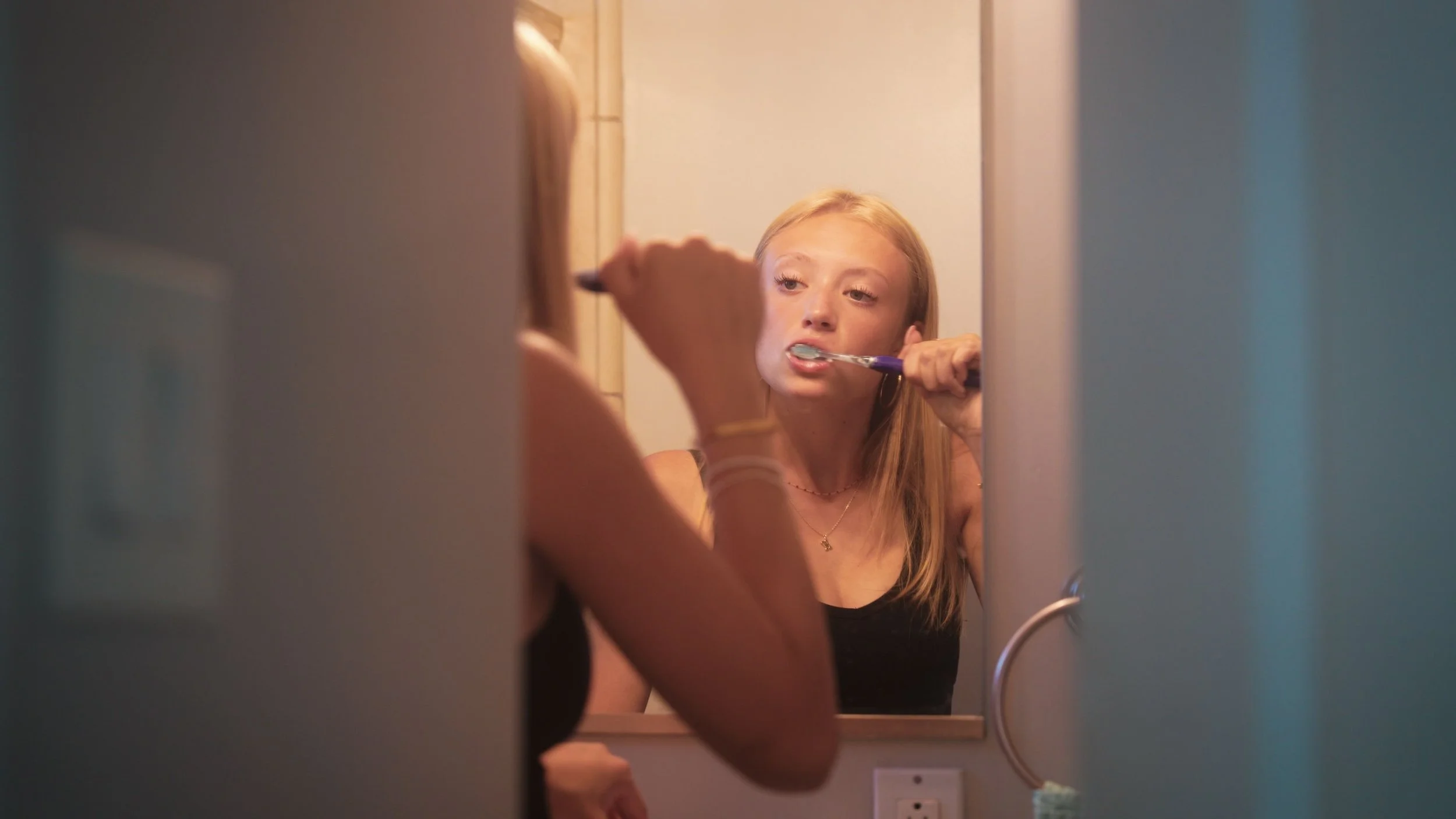 A young girl with long blonde hair brushing her teeth while looking into a mirror in a bathroom.