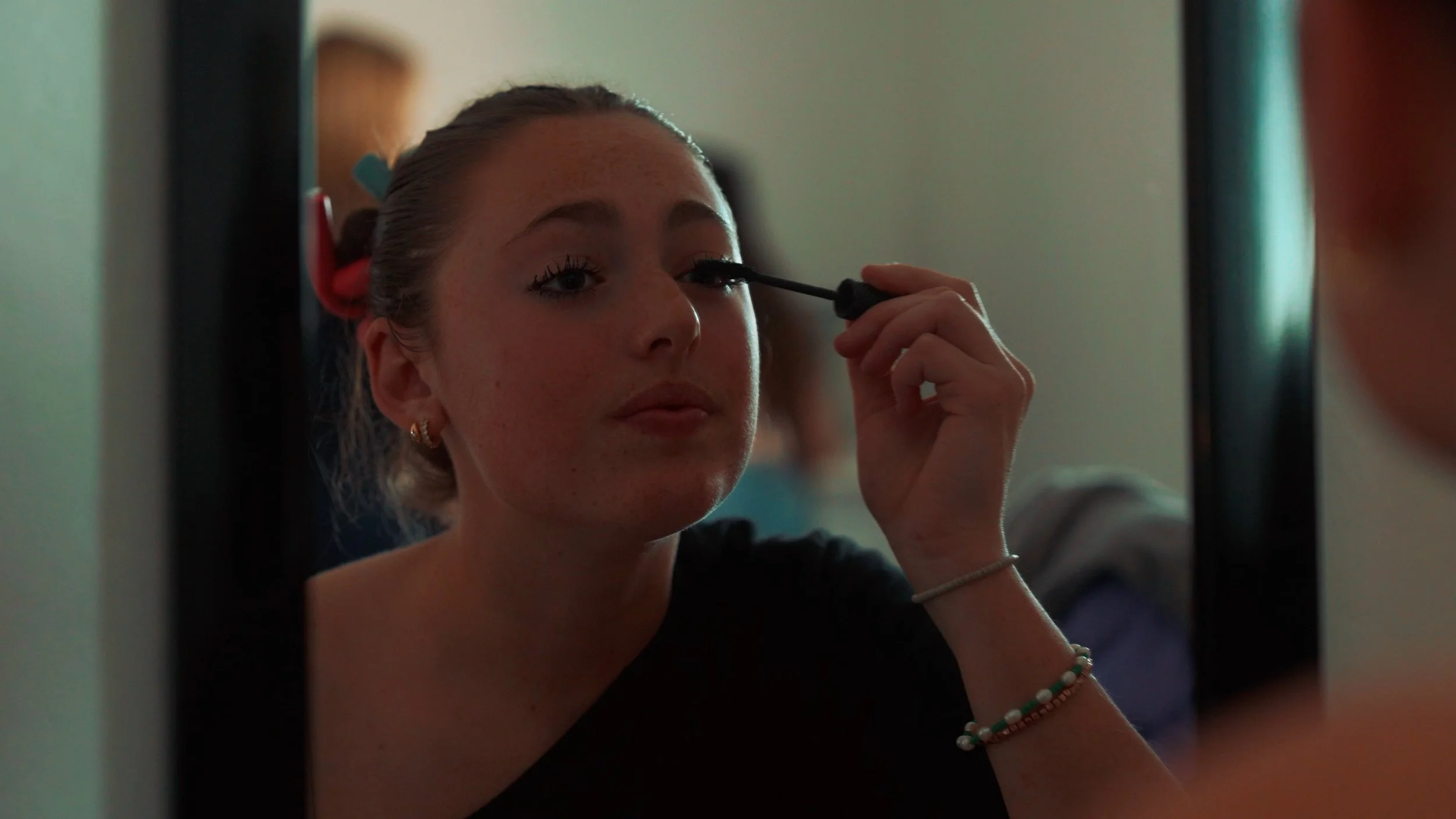 A woman with hair rollers applying mascara in front of a mirror.