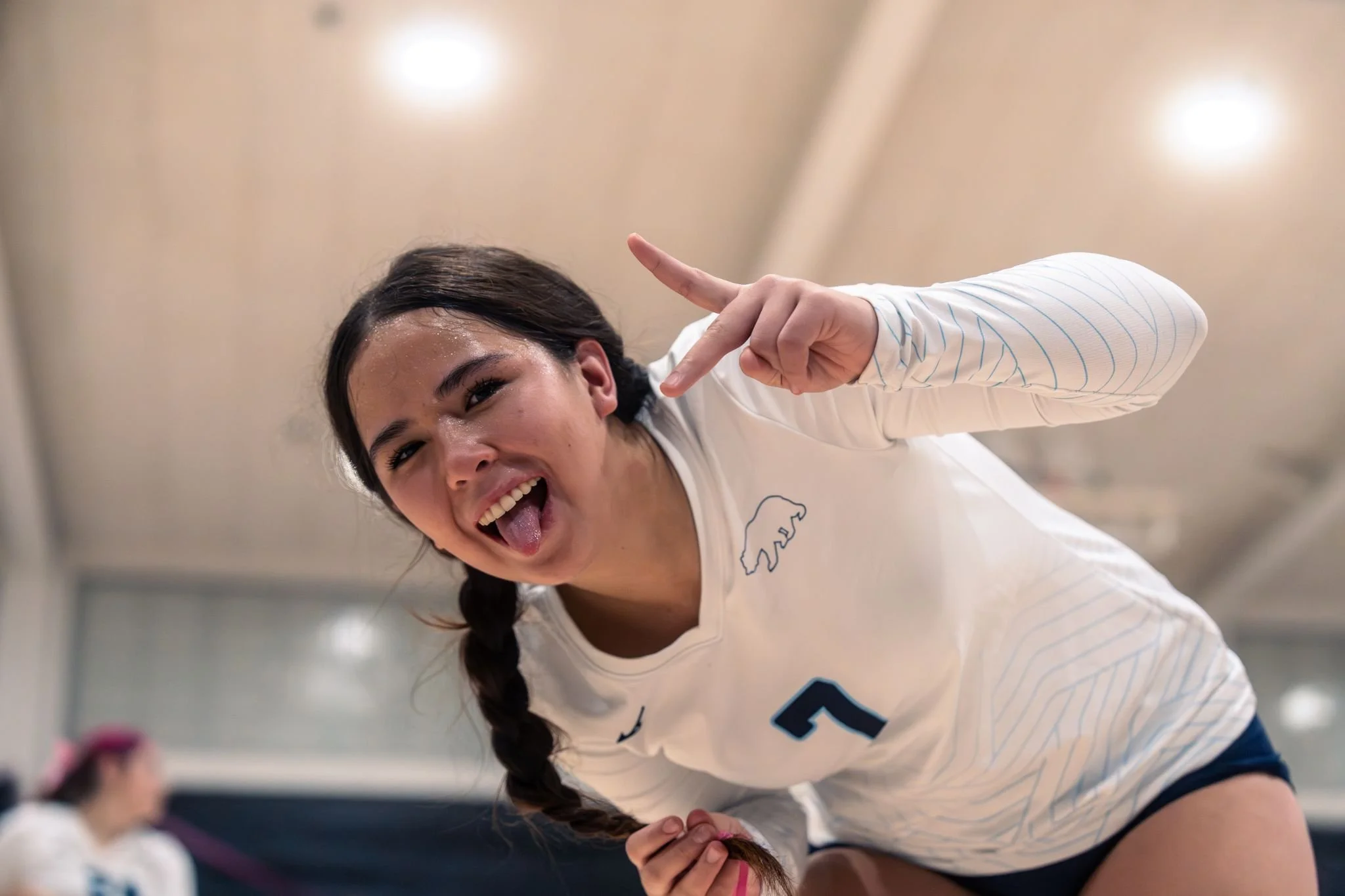A young woman with long dark hair tied in a braid is smiling, sticking out her tongue, and making a peace sign with her right hand.