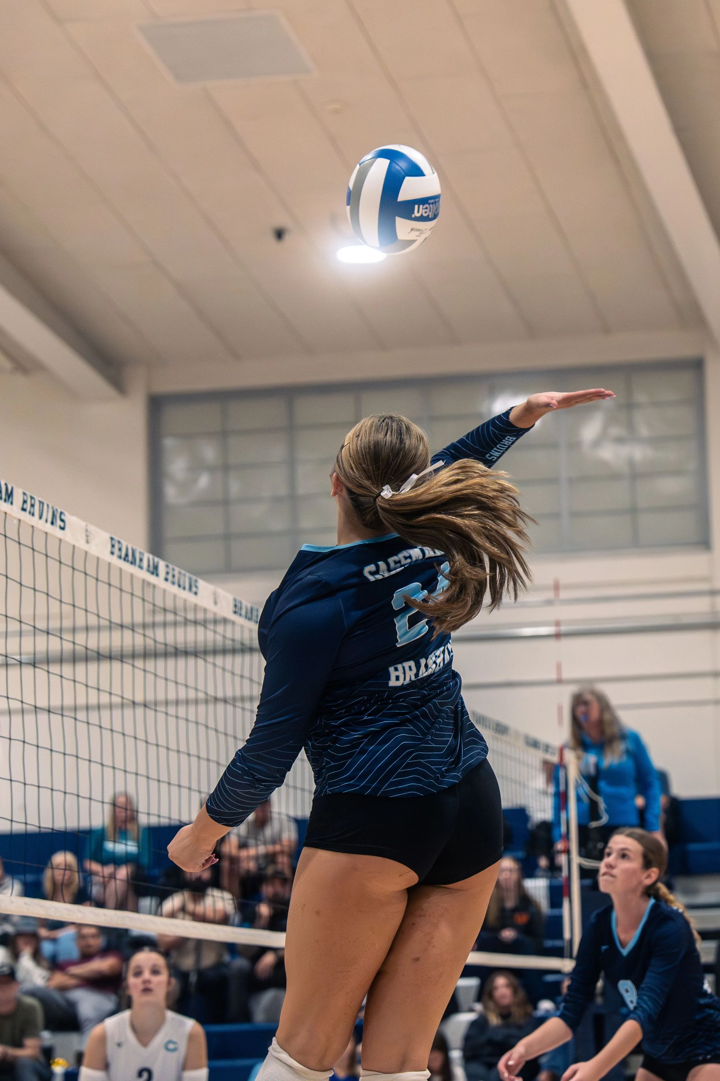 A female volleyball player in a blue uniform spiking a volleyball over the net during a match with spectators watching in the background.
