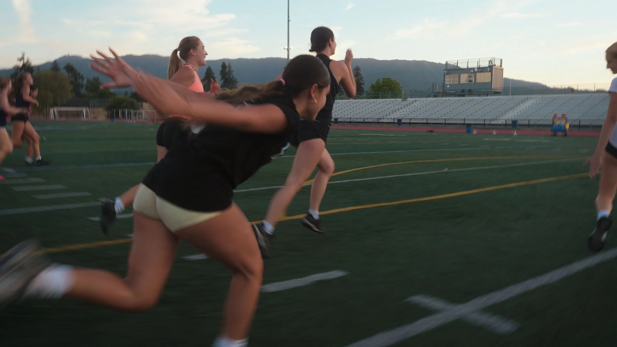 Women in athletic clothes running on a track in a stadium during sunset.