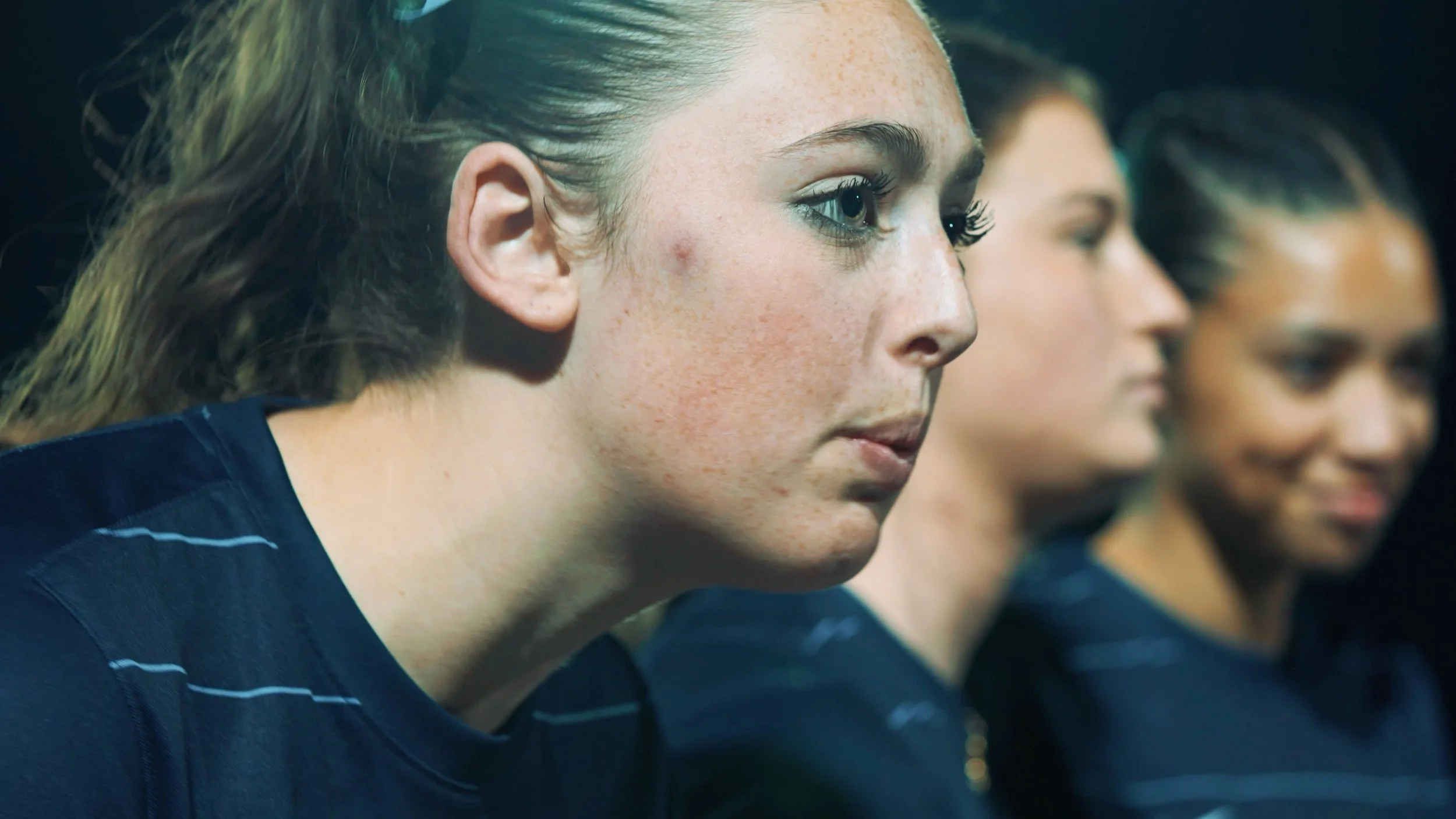 Close-up of a young woman with light skin, freckles, and blue eyes, wearing a dark top with white stripes, sitting in a row with other women, all looking attentively forward.