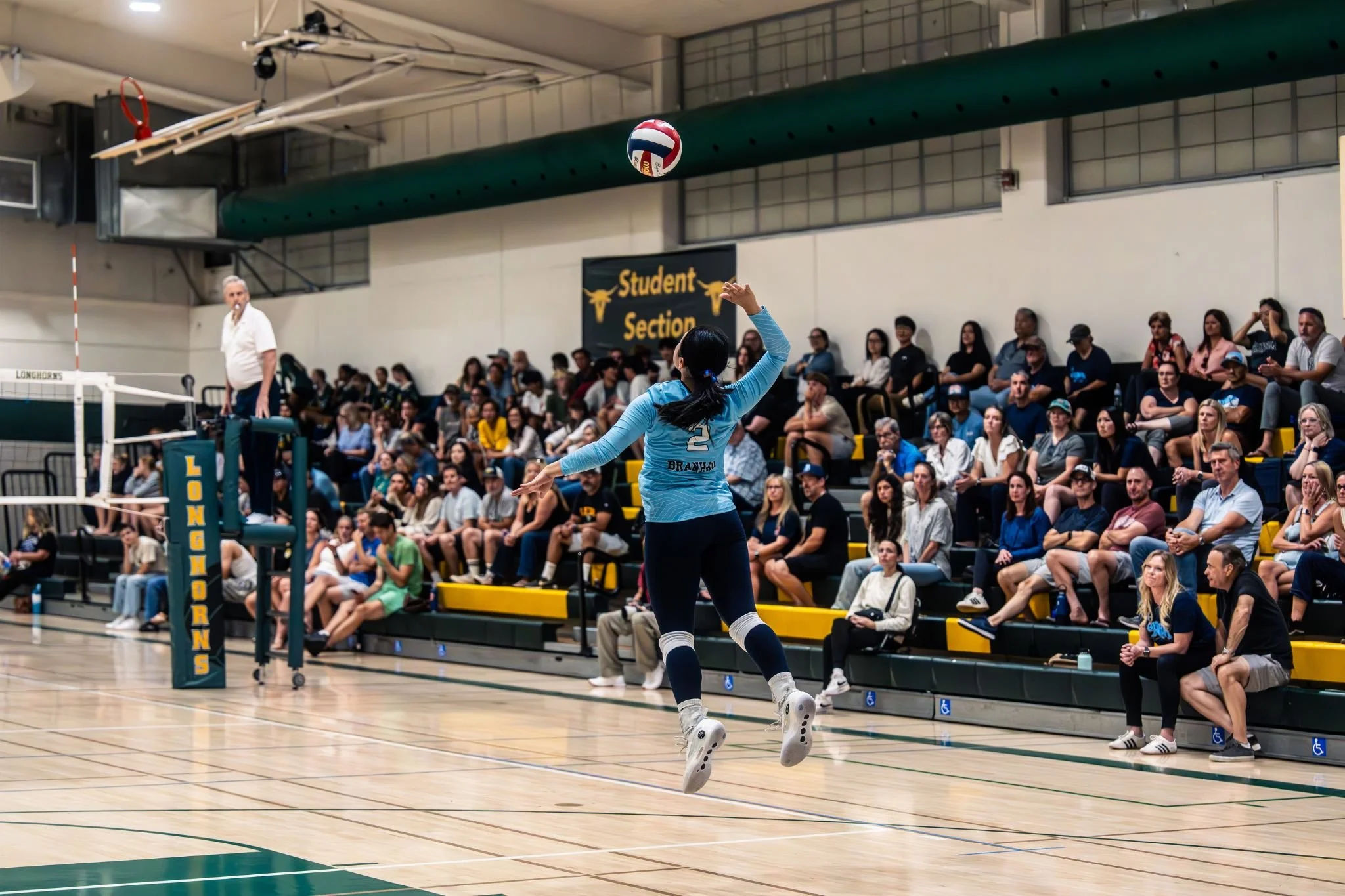 A female volleyball player in a blue jersey with the number 2 on the back is jumping to serve the volleyball during a match in a gymnasium. She is positioned near the net with her arm extended, preparing to hit the ball. Spectators are seated in the 