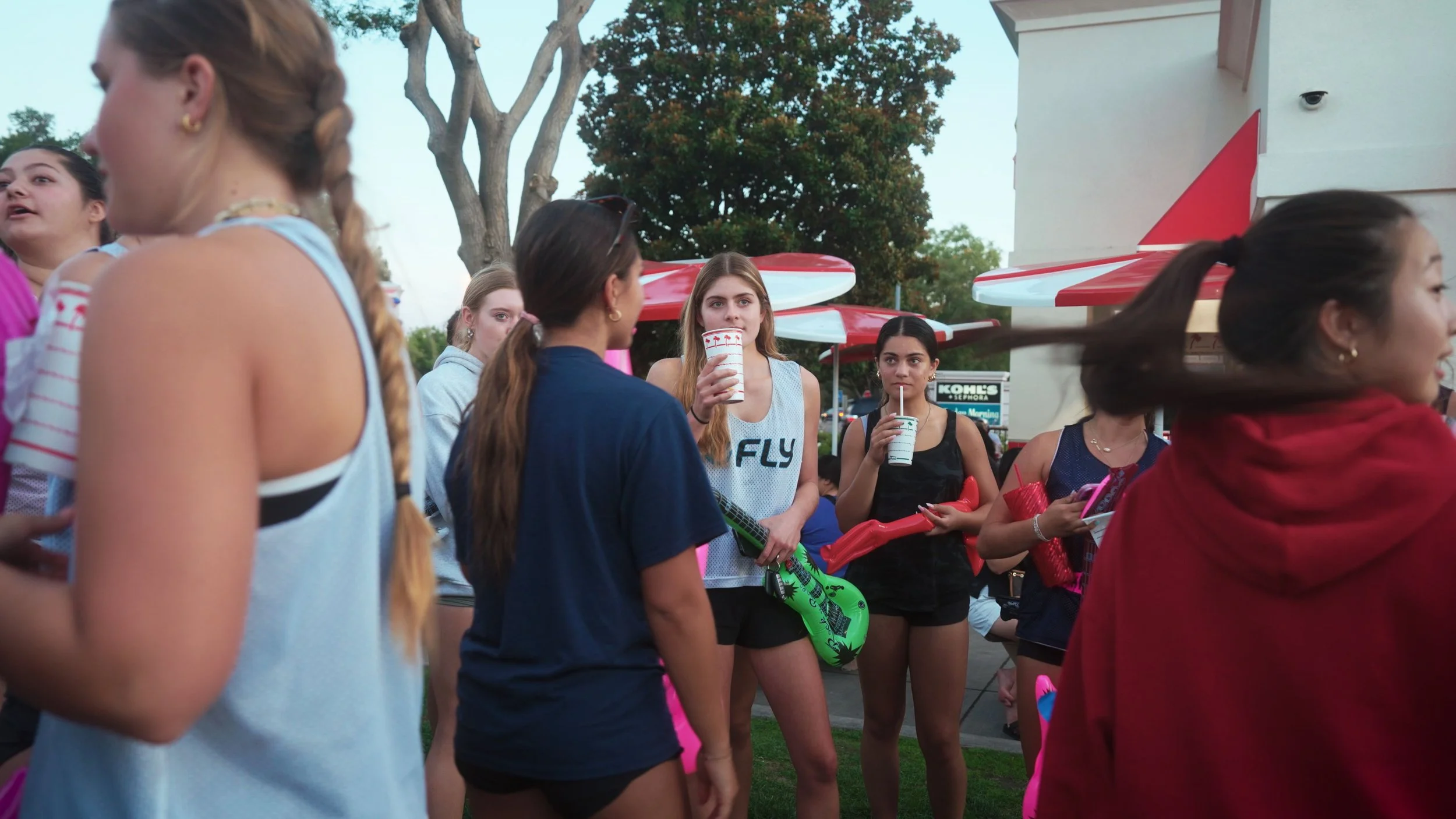 A group of teenage girls gathered outdoors on a sidewalk, some holding drinks with straws, amidst red and white umbrellas and trees, suggesting a casual social setting.