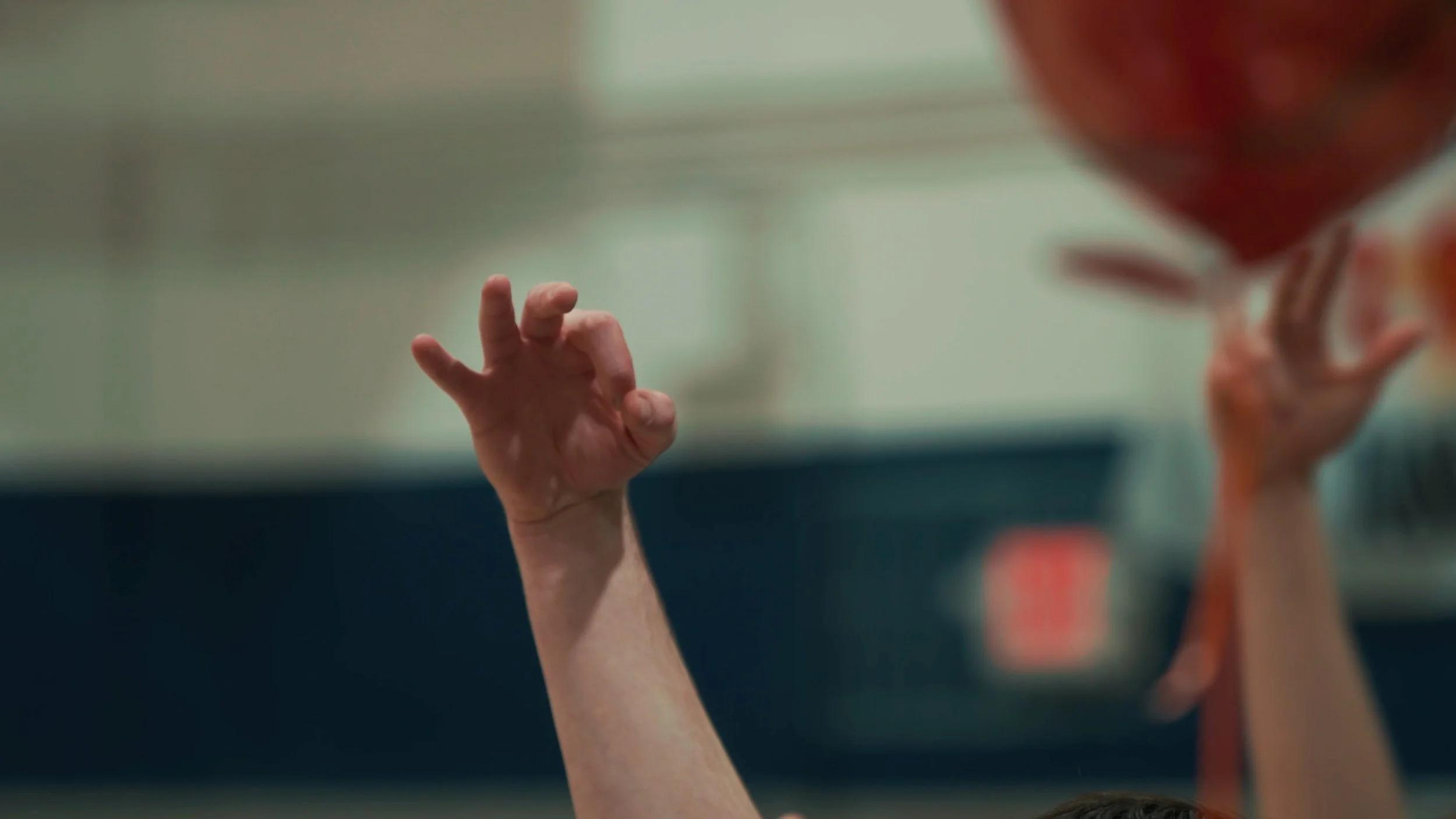 A person reaching up with their hand, holding a red basketball while another person in the background also reaches for it.