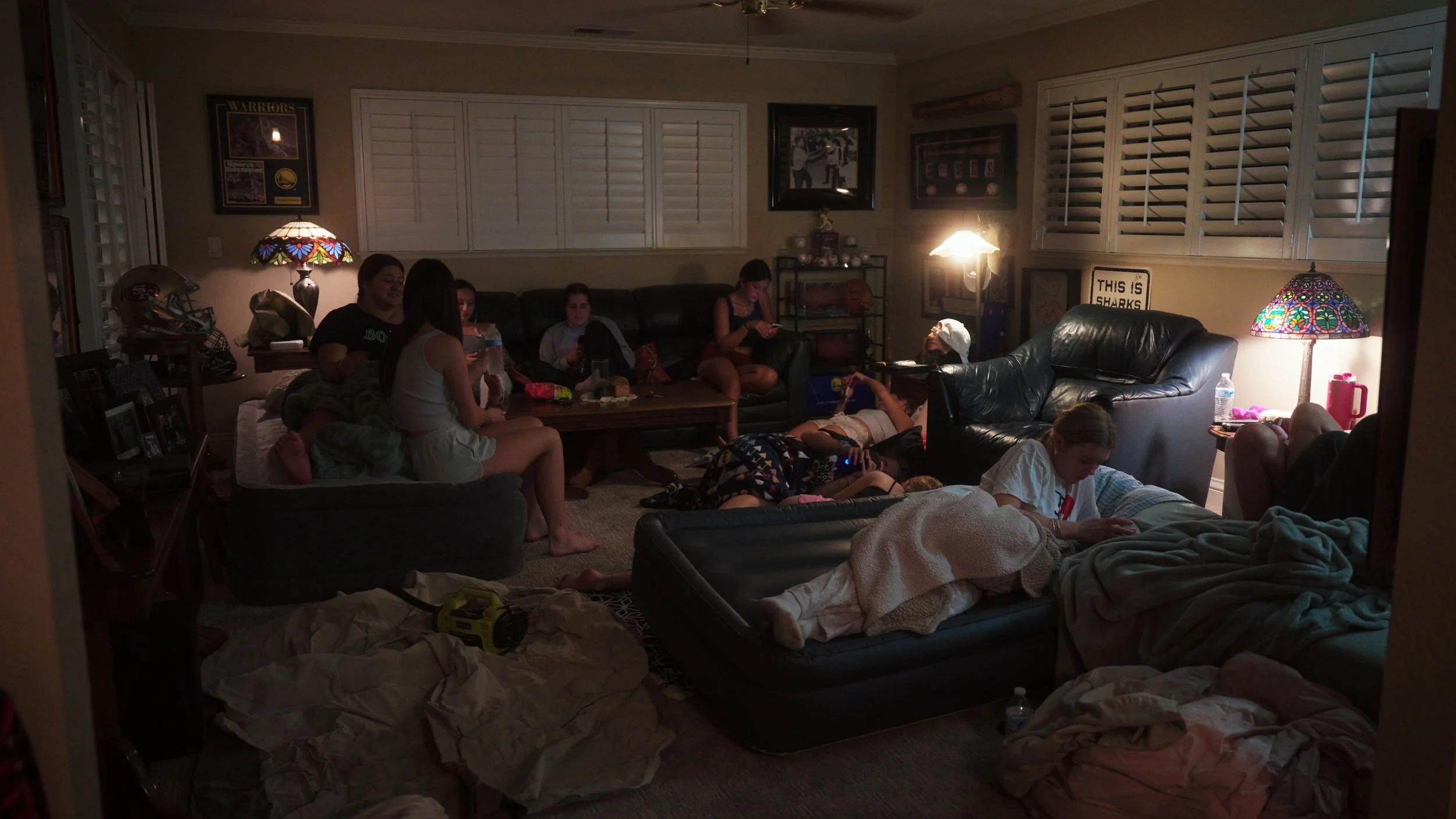 A dimly lit living room filled with people, mostly children and teenagers, lounging on couches and the floor, some using phones or looking around. There are lamps with stained glass shades, a sign that reads 'This is Sharks,' and various framed pictu