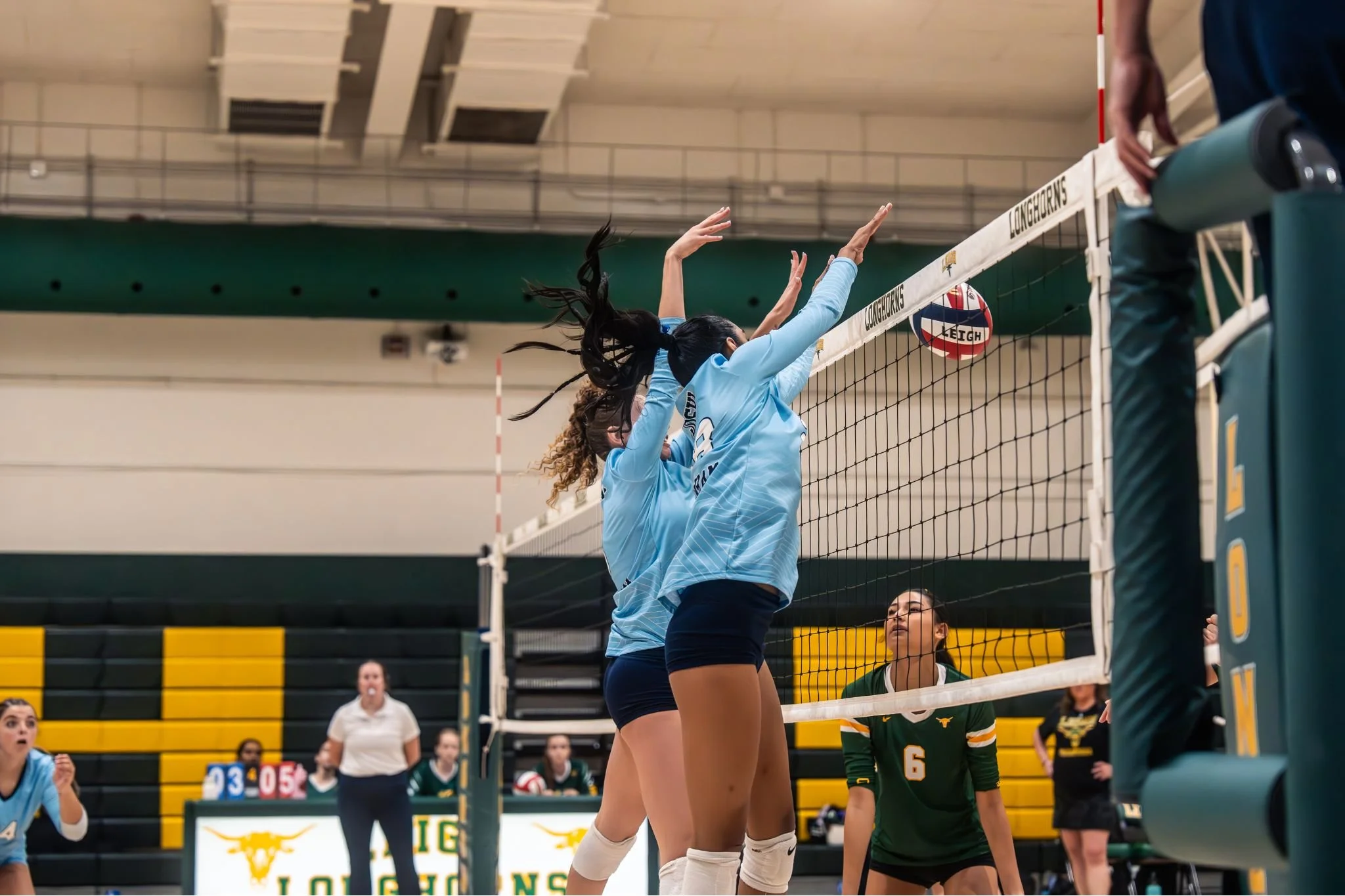 Two volleyball players jump at the net during a game, with one in a light blue jersey and black shorts and the other in a green and yellow jersey. Spectators and officials are in the background.