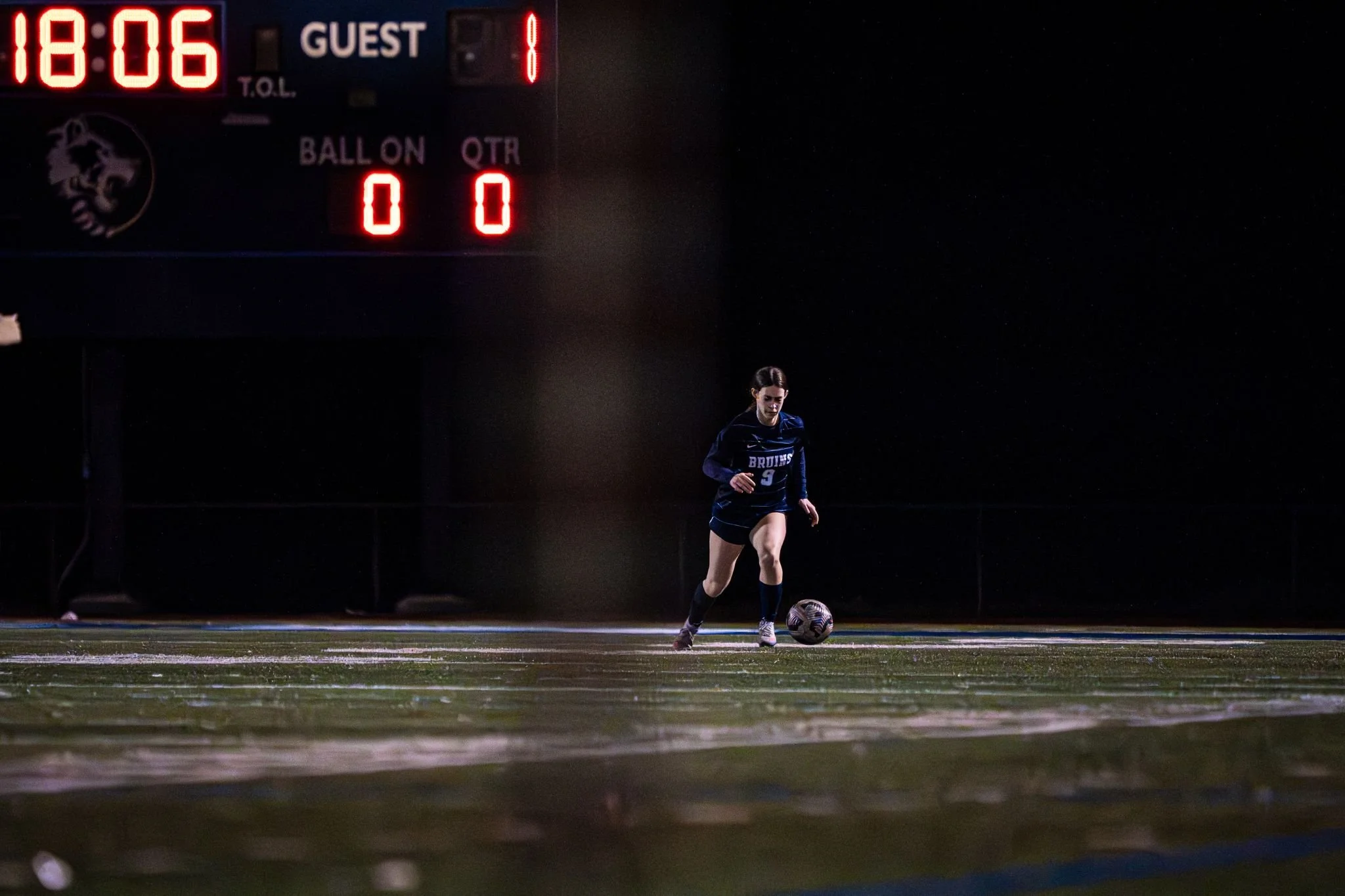 A female soccer player in a navy uniform with the number 9 on her jersey, dribbling a soccer ball on a football field at night under stadium lights. A scoreboard in the background shows the time 18:06 and the score as 0-1.