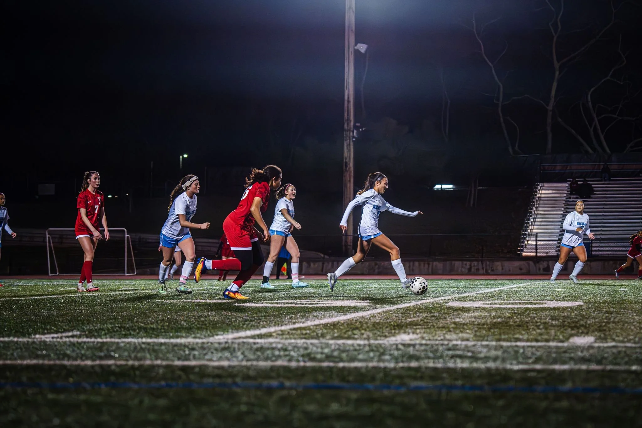 A women's soccer game is underway on a well-lit field at night. Several players, wearing red and white jerseys, are running towards the ball, with one player preparing to kick it. Bleachers are visible in the background, and the field's artificial tu