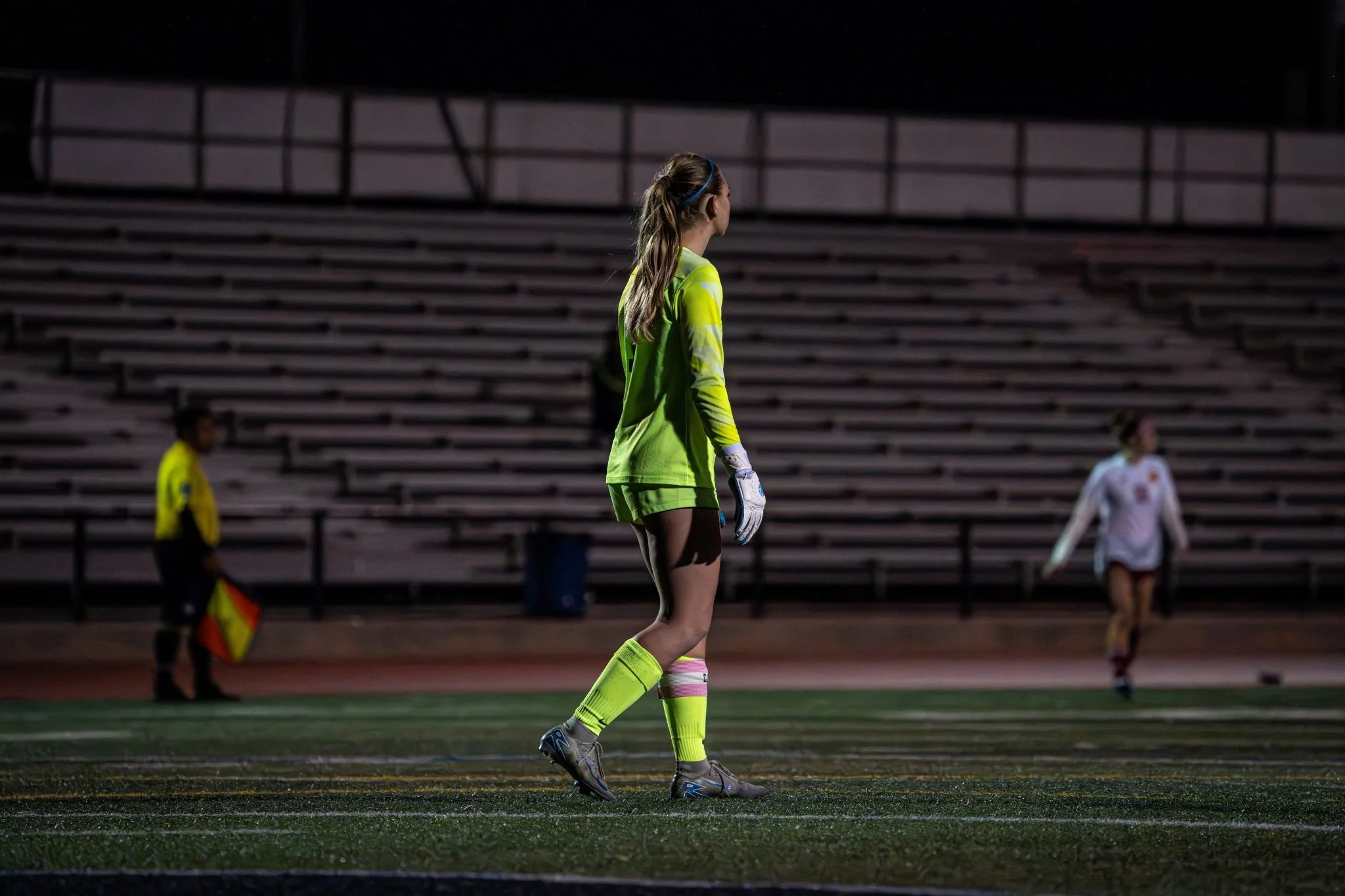 Female soccer goalie standing on the field at night, wearing a yellow goalkeeper jersey and gloves, with other players in the background.