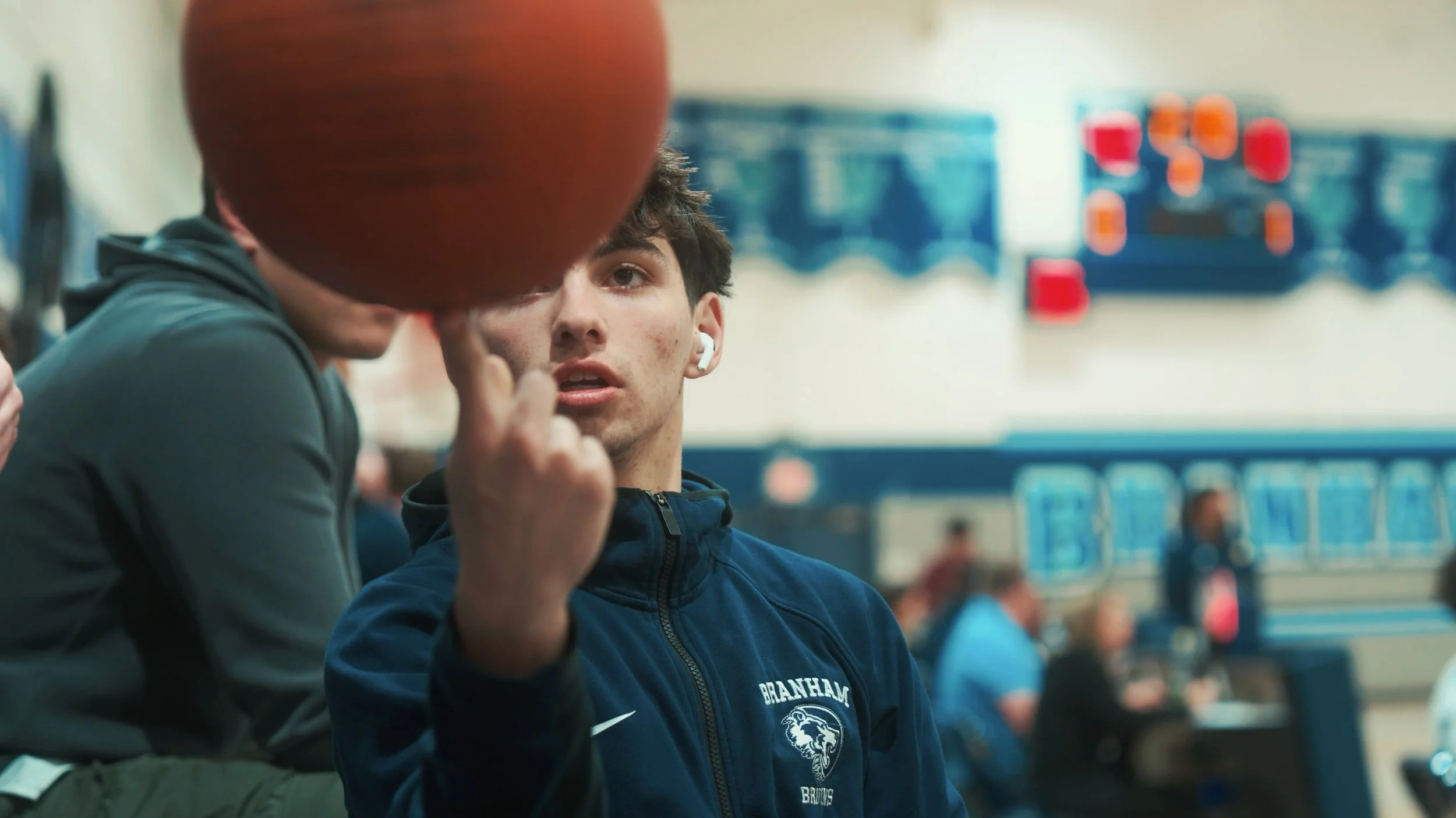 A young man with brown hair, wearing a dark blue jacket, is looking at another person who is spinning a basketball on their finger. The background suggests they are in a gymnasium with a basketball scoreboard and blue bleachers.
