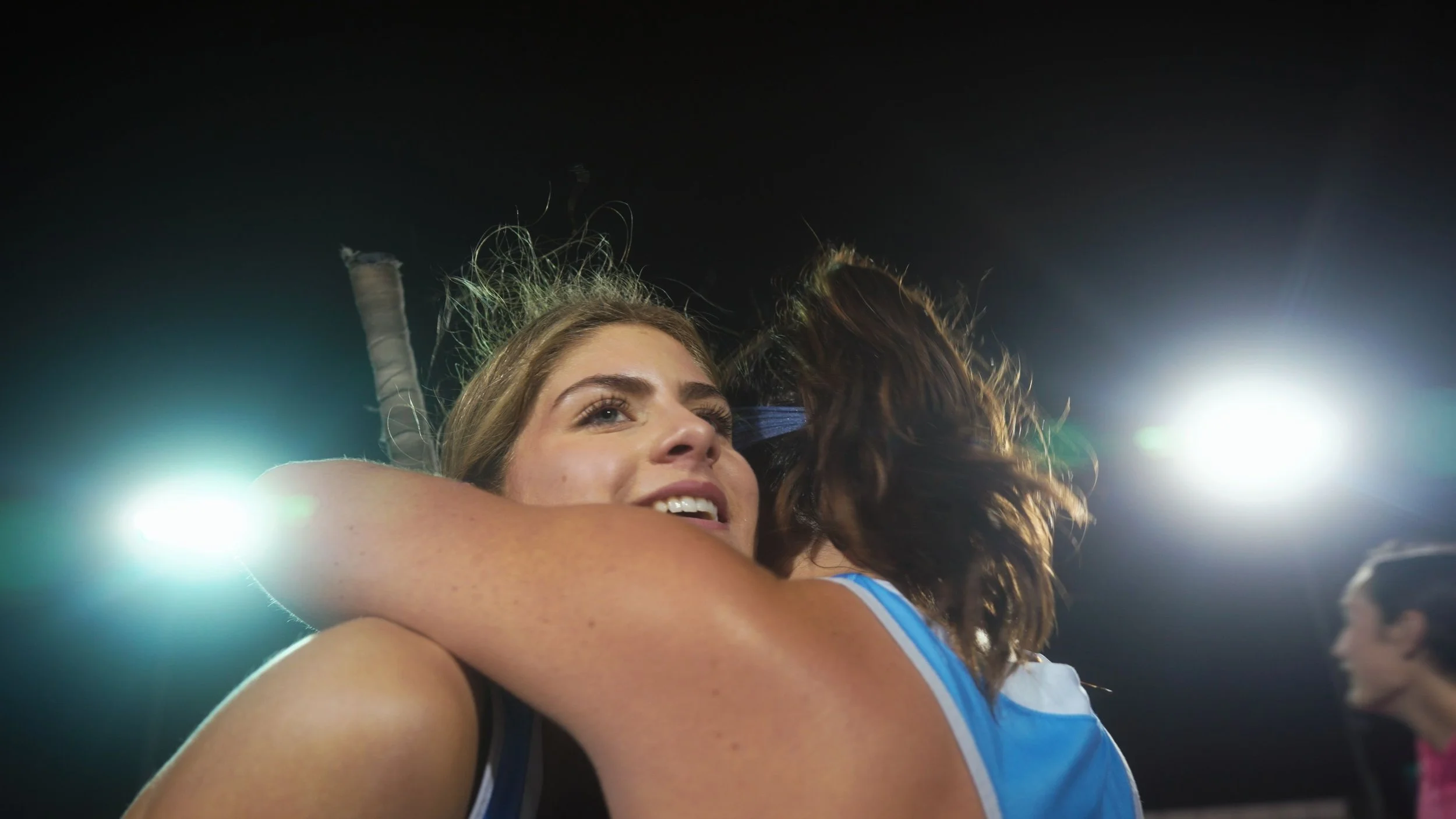 Two women hugging at a sports stadium, with bright stadium lights in the background.